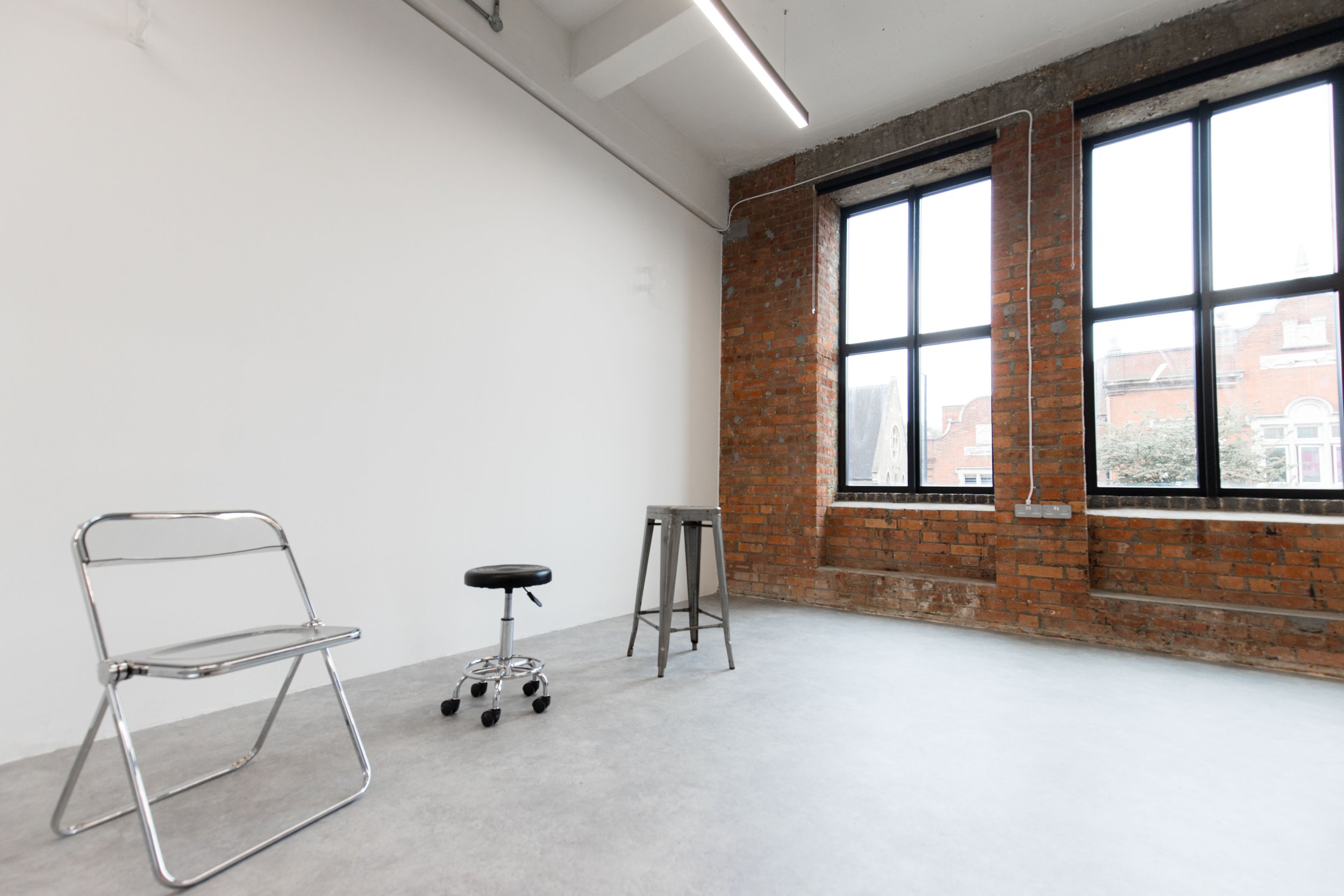 An unfinished interior space with large windows, a white wall, exposed brick wall, and construction tools including a stool, a ladder, and a rolling cart.