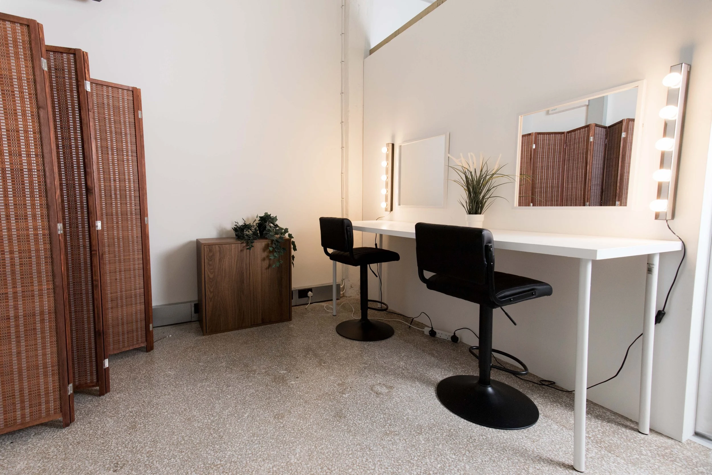 A room with a white wall and a mirror with makeup lights, a white desk with a large potted plant, two black chairs, a wooden cabinet, and a wooden room divider.