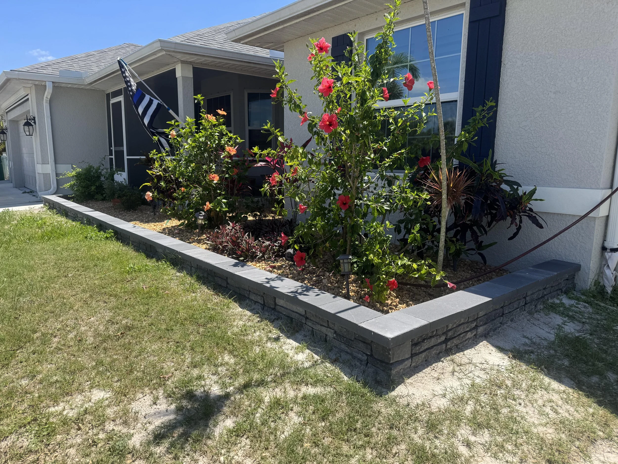 A house with a porch, garden, and flower bed with pink hibiscus flowers, small plants, and garden lights.