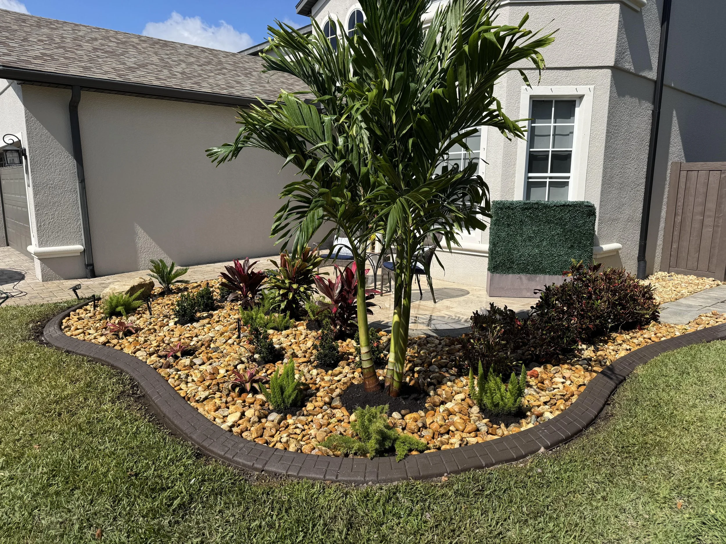 A landscaped front yard with a small garden bed filled with various plants and surrounded by a black plastic border. There is a tall palm plant in the center with other smaller plants around it, and a house with white and gray exterior walls, white-framed windows, a shingled roof, and a small patio with a chair and pink flowers in the background.