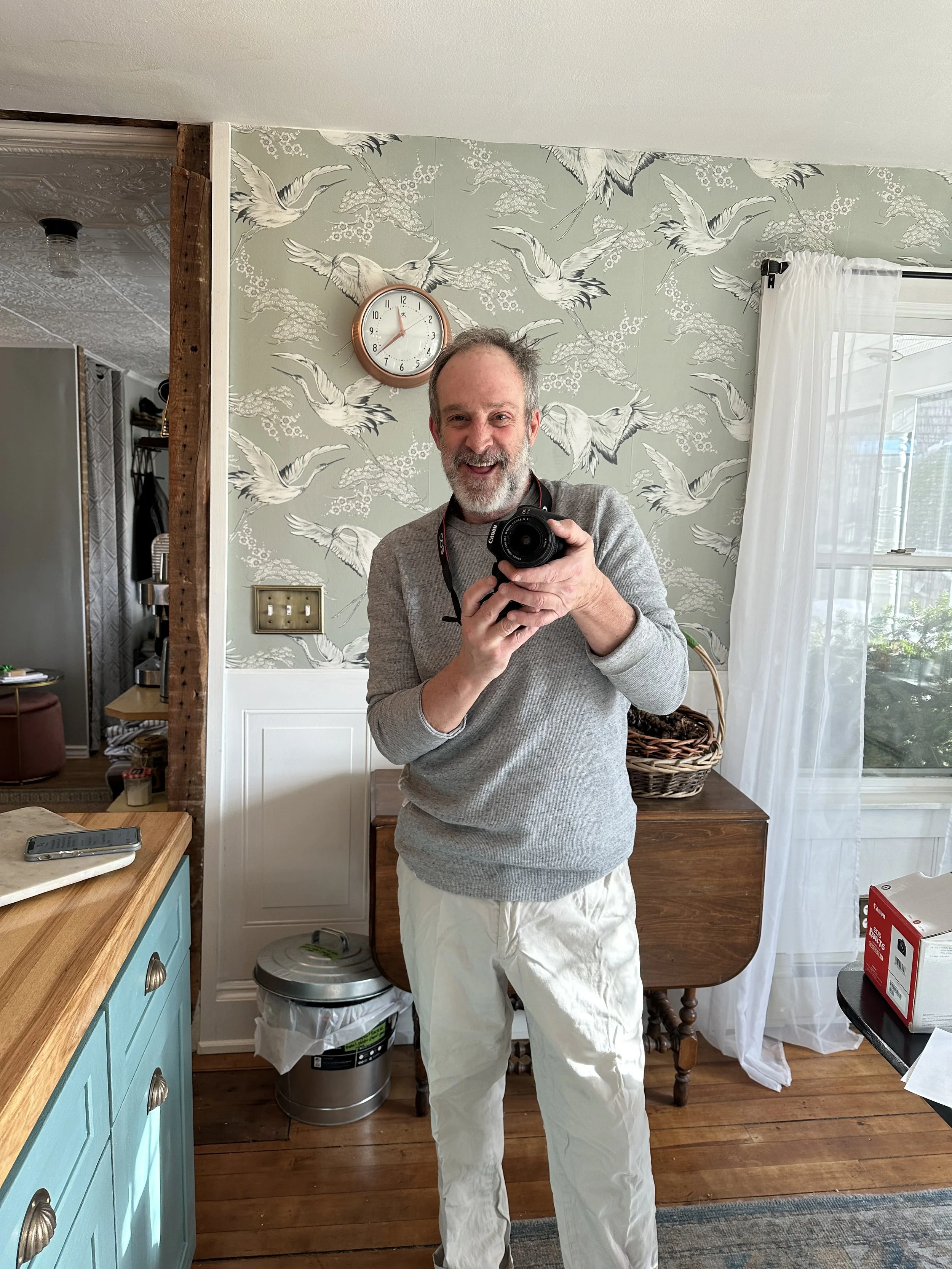 A middle-aged man with gray hair and beard taking a picture in his kitchen, wearing a gray sweater and white pants.