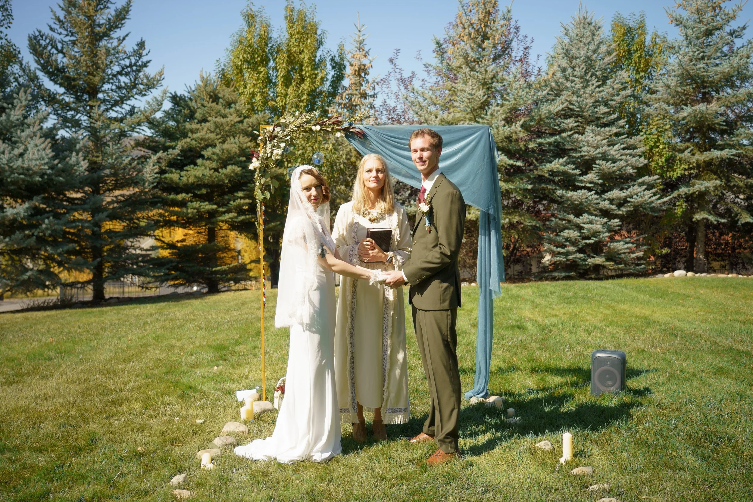 A couple getting married outdoors with a female officiant, standing under a decorated arch made with blue fabric and greenery, surrounded by trees.
