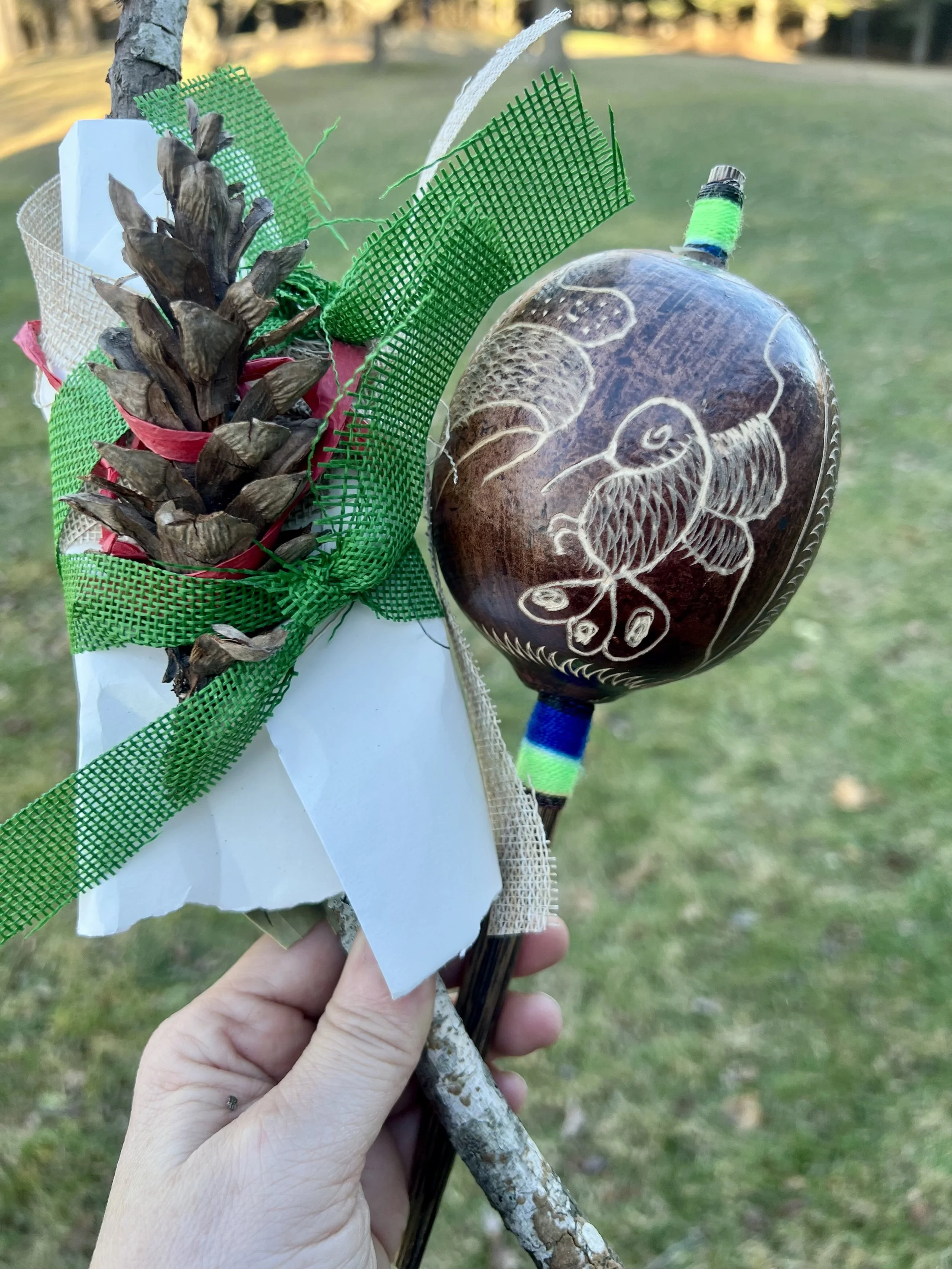A person holding a decorated stick with natural and craft materials, including pinecones, green mesh, and carved dark wood, with an outdoor grassy area in the background.
