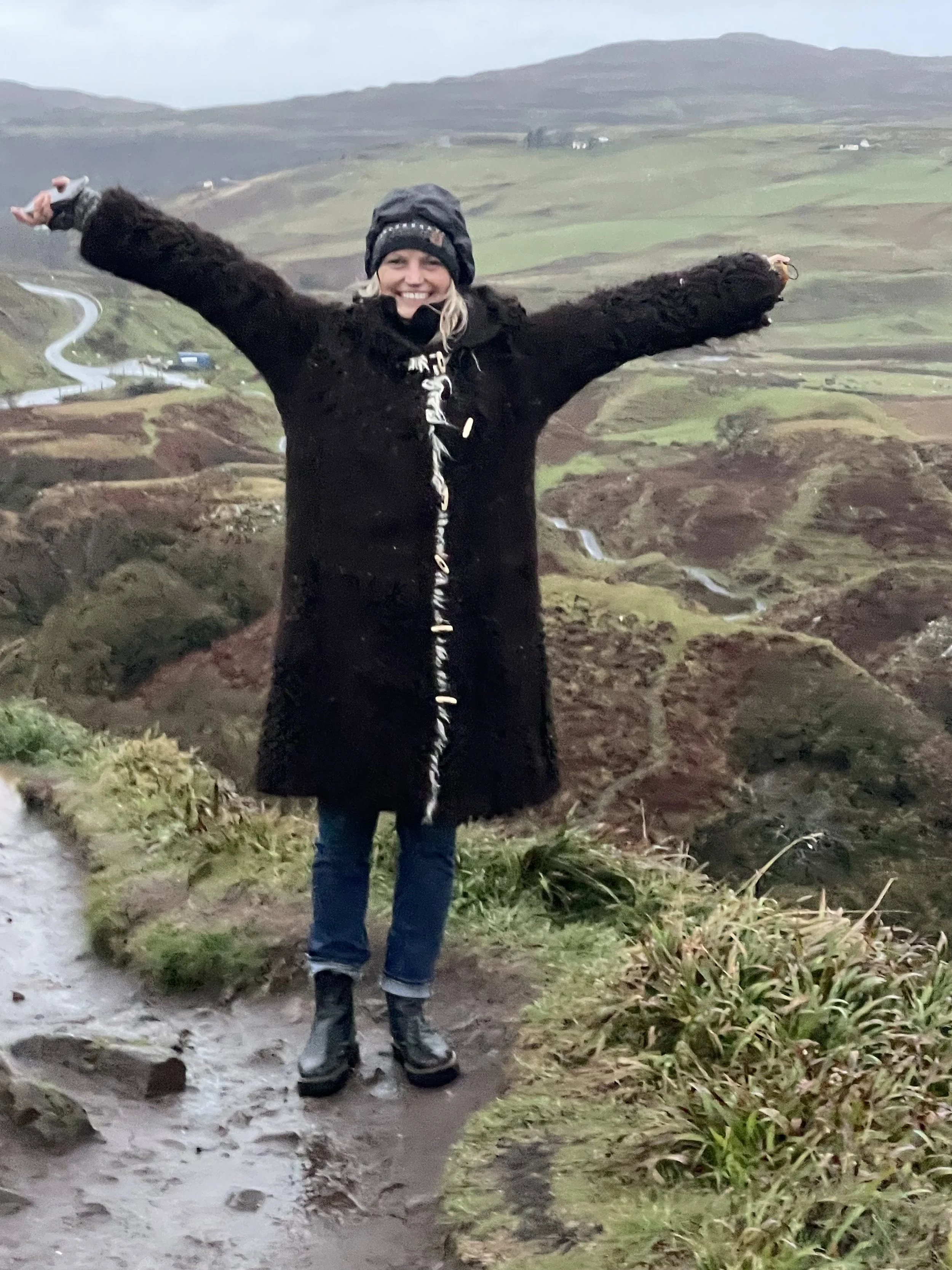 Woman standing outdoors on a muddy trail, arms raised, with rolling green hills and winding roads in the background.