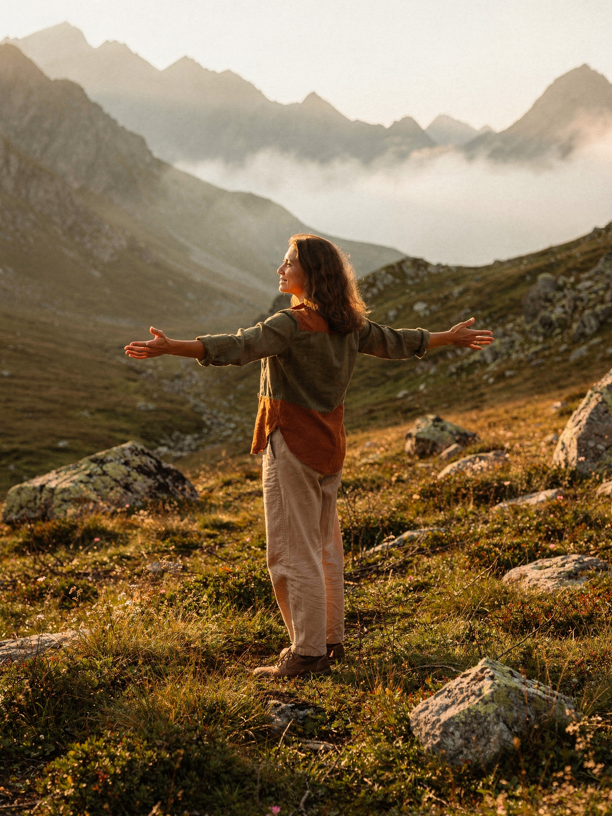 Mujer de espaldas con brazos abiertos en un paisaje montañoso al atardecer.