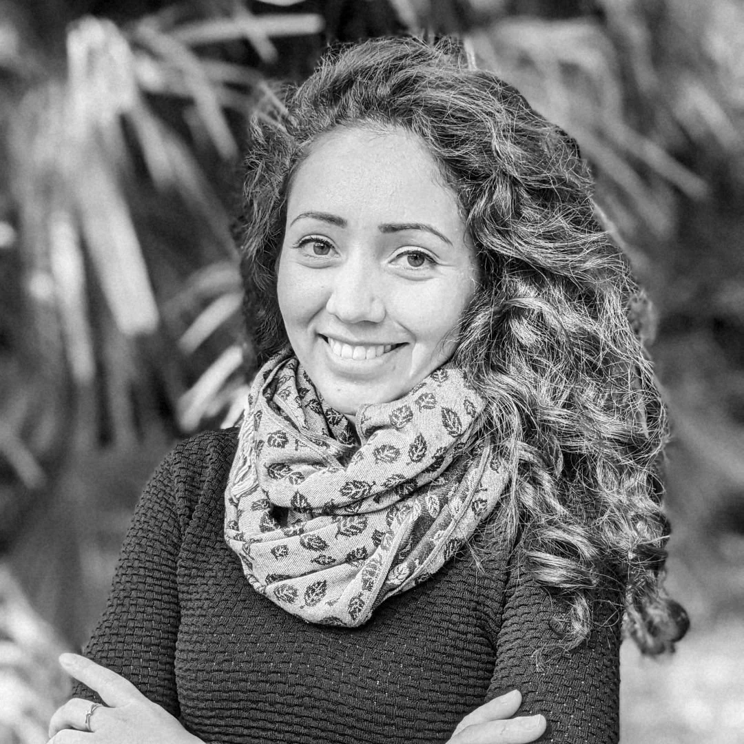 Retrato de una mujer sonriendo, con cabello rizado, usando un suéter oscuro y un bufanda de estampado floral, en un entorno natural con plantas en el fondo.