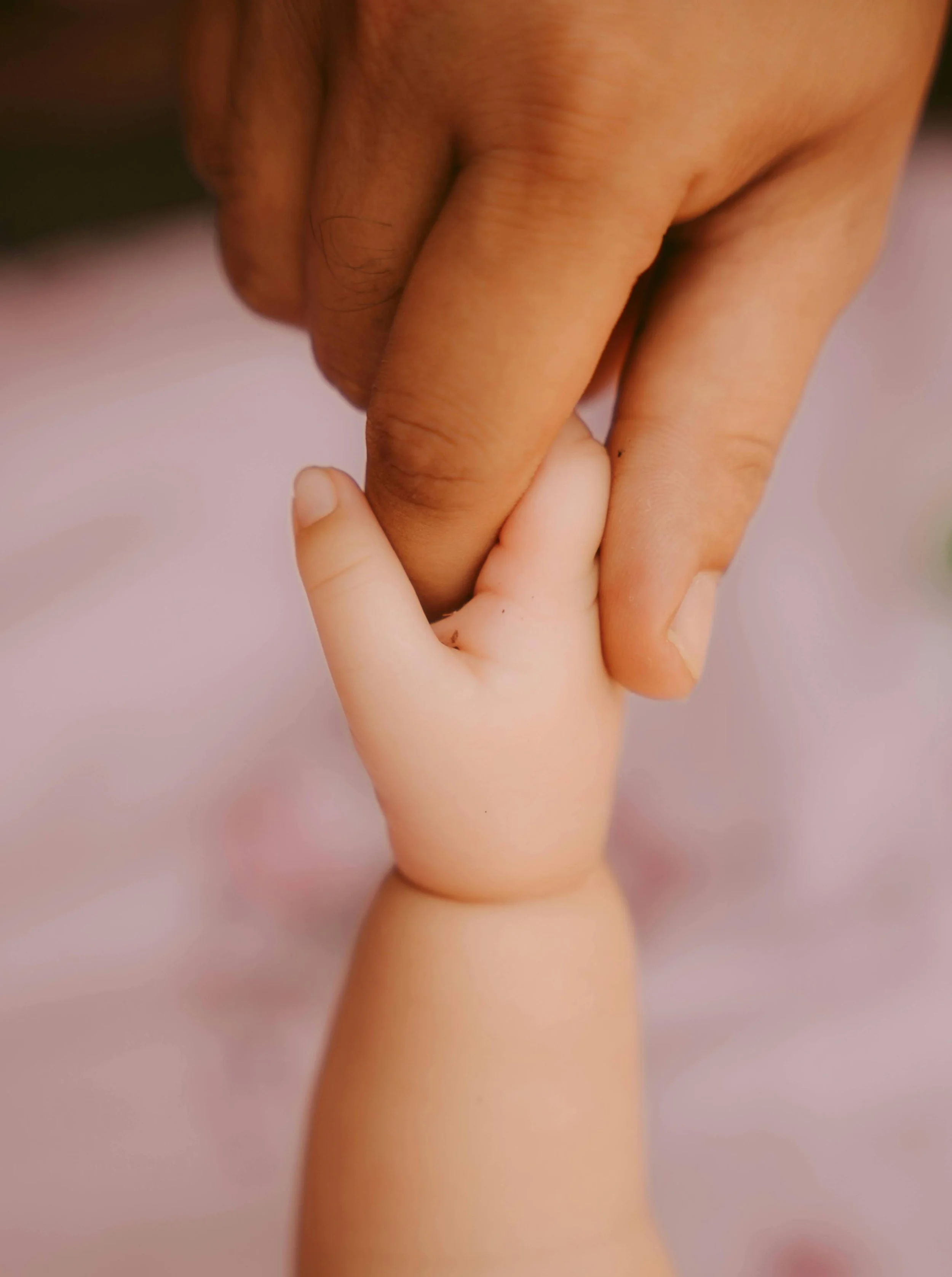Infant hand holds parent's finger with soft pink background.