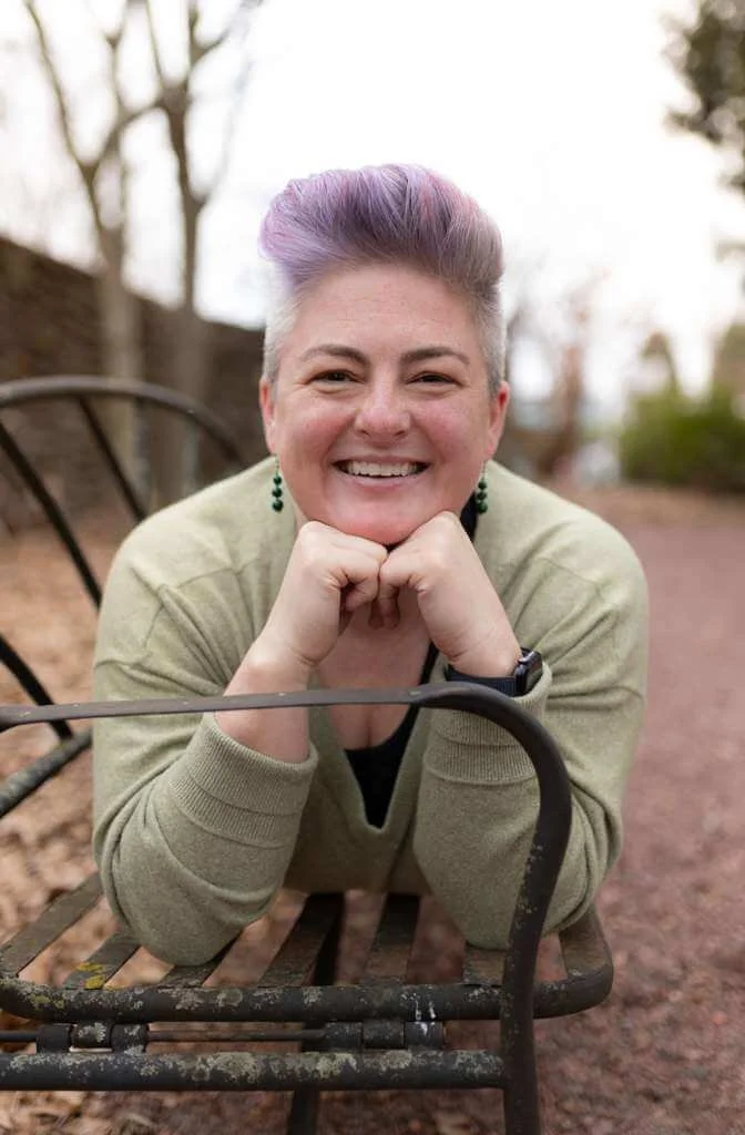 Postpartum doula Wendy Black smiles while laying on a park bench with her chin resting on her hands and wearing a light green sweater.