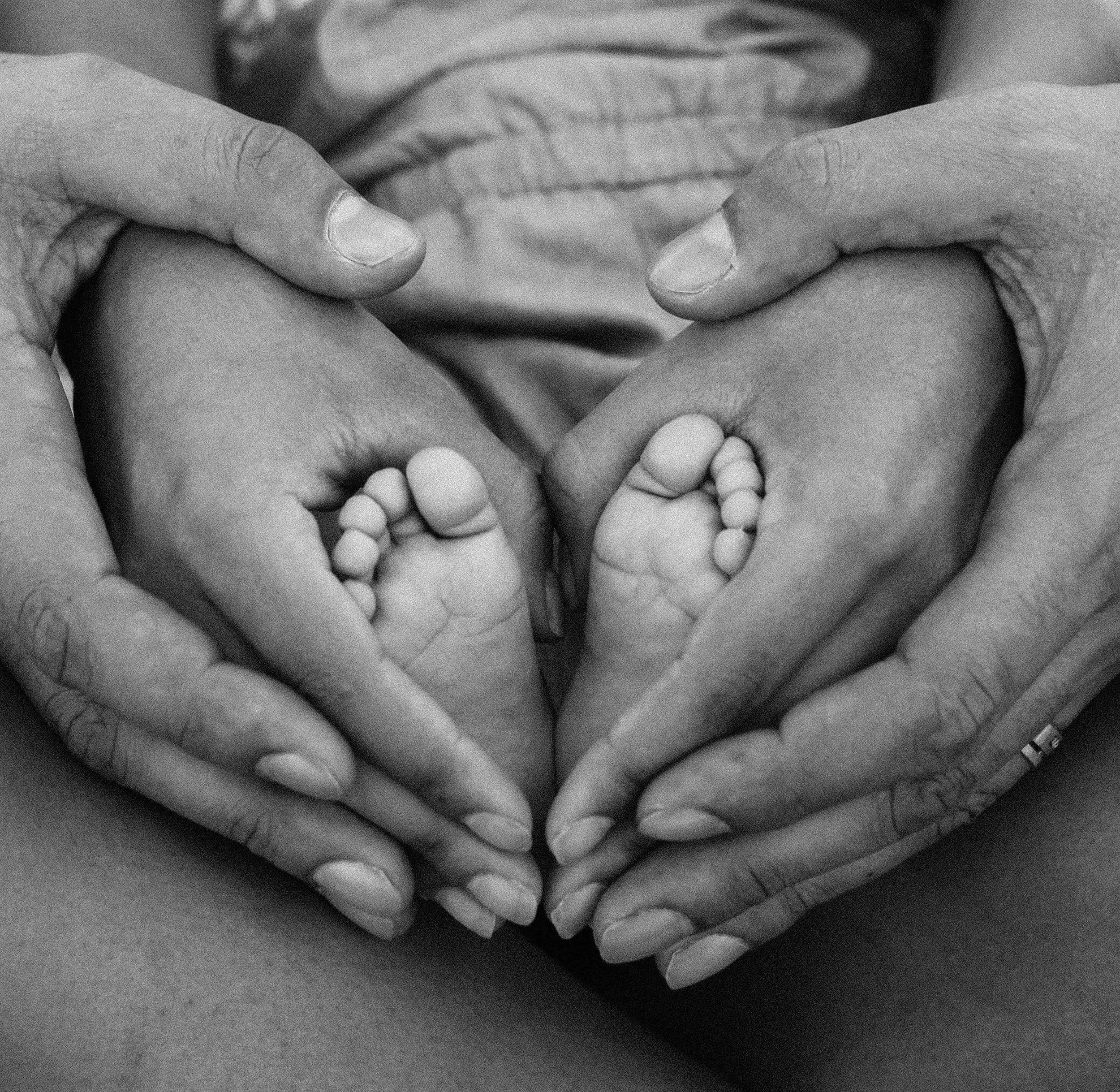 Black and white image of an infant's feet cradled by their parents' hands in the shape of a heart.
