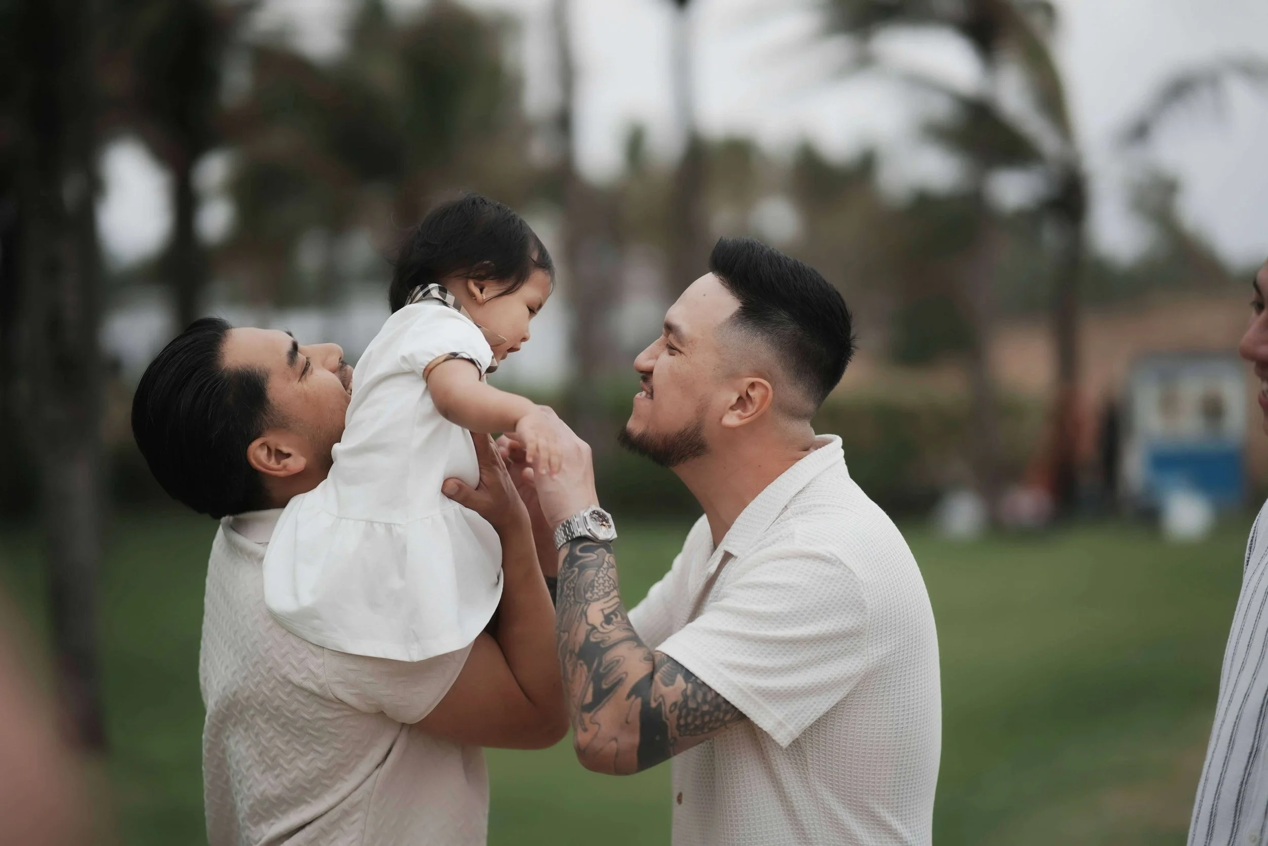 One dad holds their infant daughter on his shoulder while the other dad smiles at her and holds her hands.