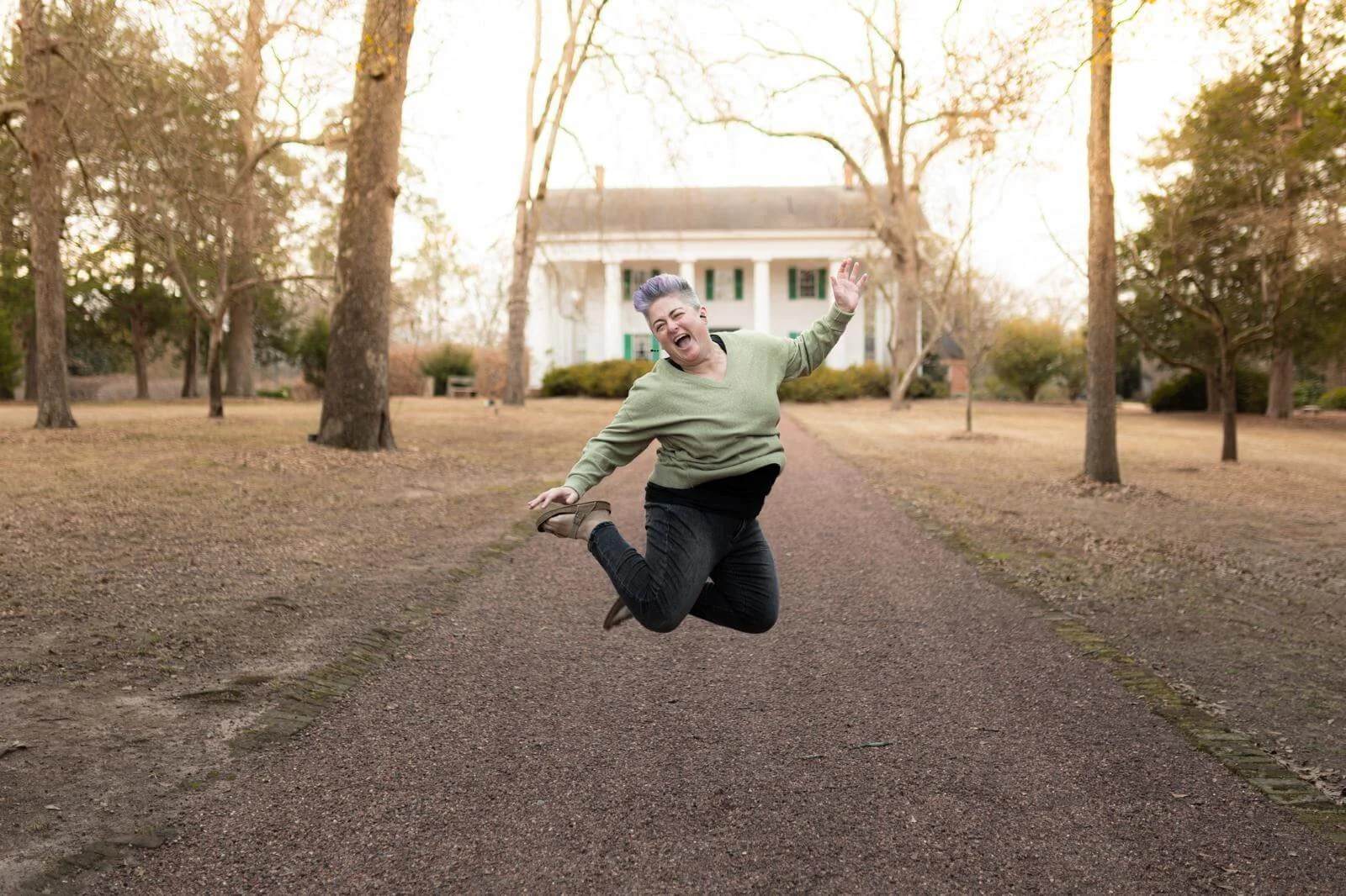 Metro Atlanta postpartum doula Wendy Black jumps in the air and smiles in front of a long driveway and a white house in the background.