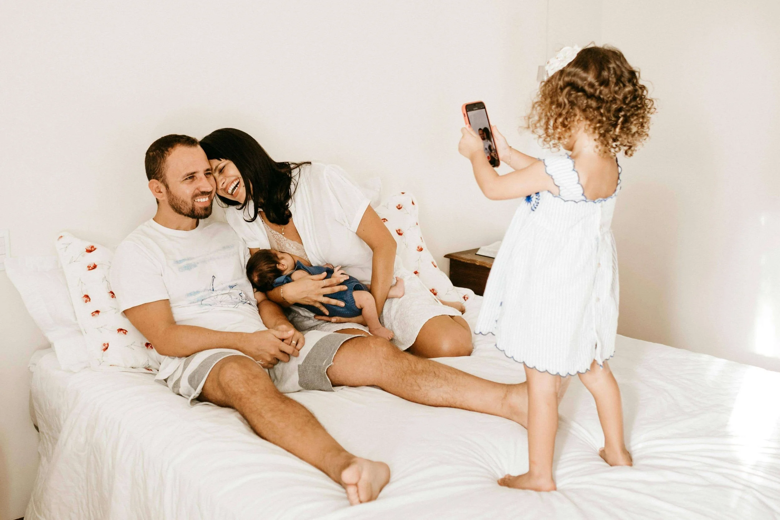 Second-time parents laugh and hold their infant in bed while their older daughter takes a photo of them.