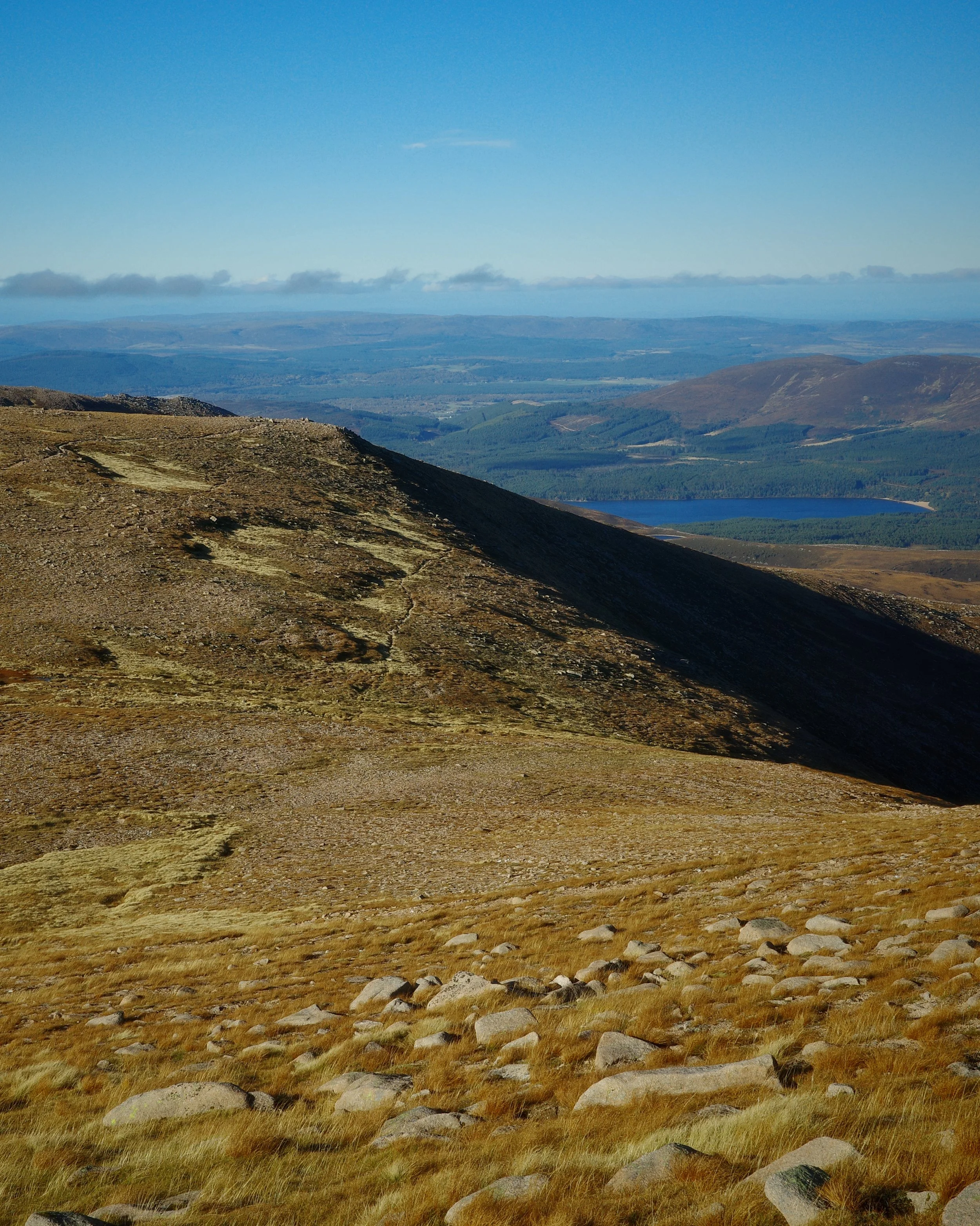 A mountain landscape with a rocky, grassy foreground, a sloped hillside, and distant lakes and hills under a partly cloudy sky.
