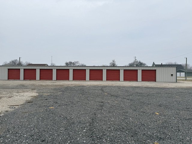 A row of storage units with red doors in an outdoor storage facility, with a gravel lot in front and overcast sky.