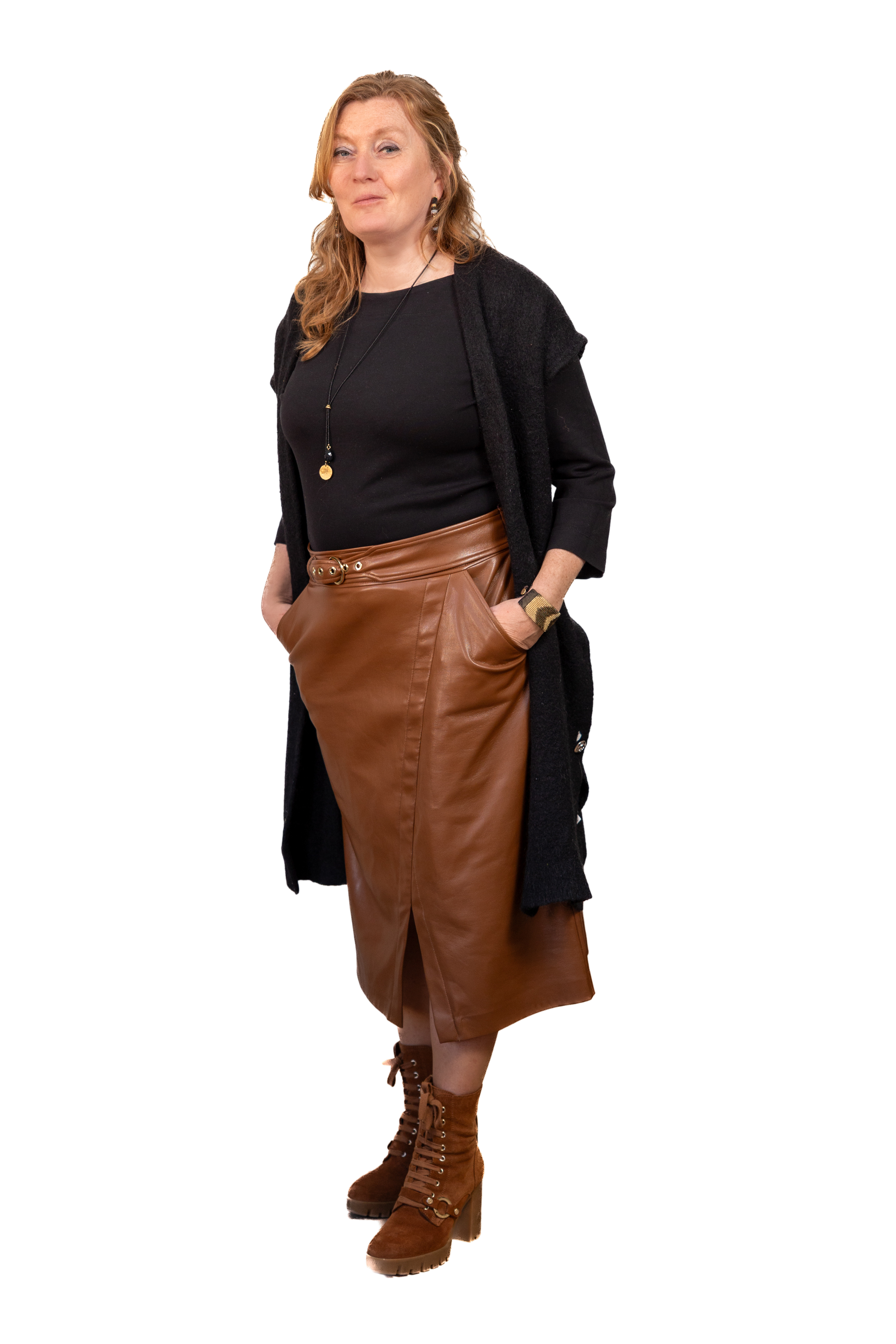 Vrouw met rode haar in een zwarte top, lange bruine leren rok, bruine laarzen en een zwart lang vest op een zwarte achtergrond.
