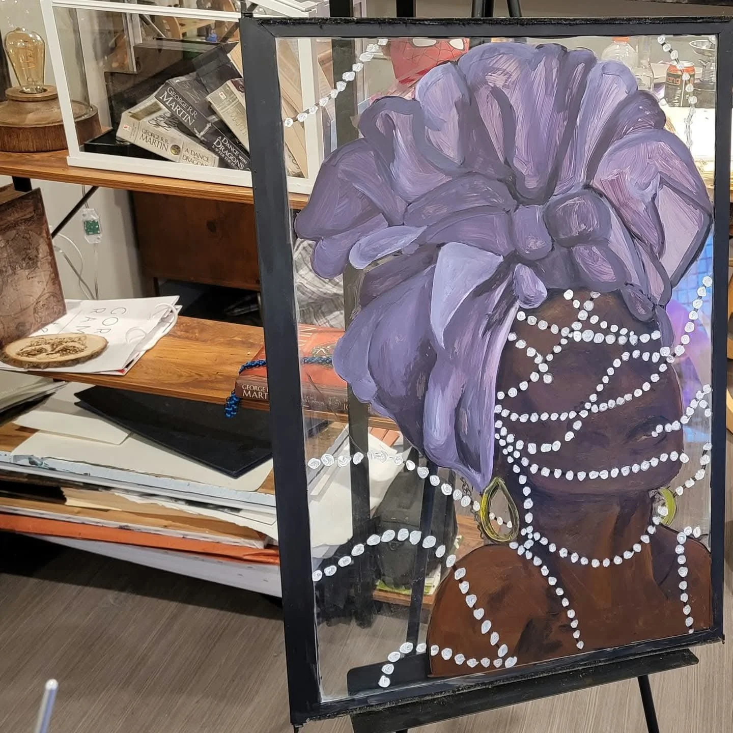 Painting of a woman wearing purple headwrap and large earrings, with decorative white beads draped across her face and shoulders, displayed on an easel in an art studio.