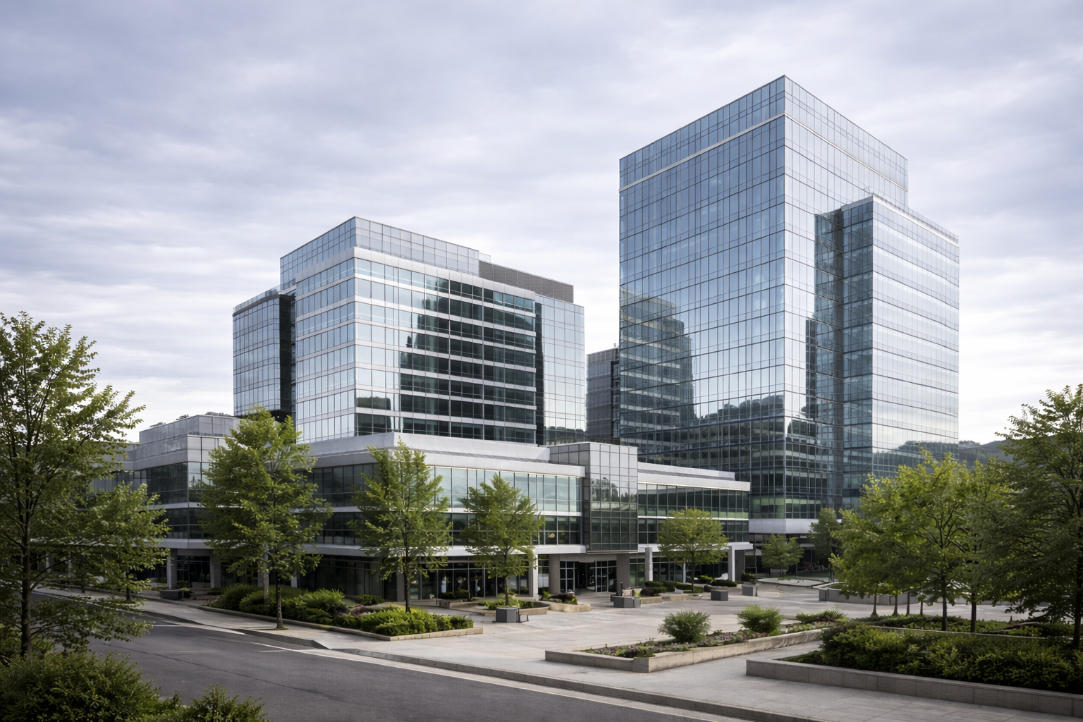 Modern glass office buildings with reflective windows, surrounded by trees and a paved plaza under a cloudy sky.