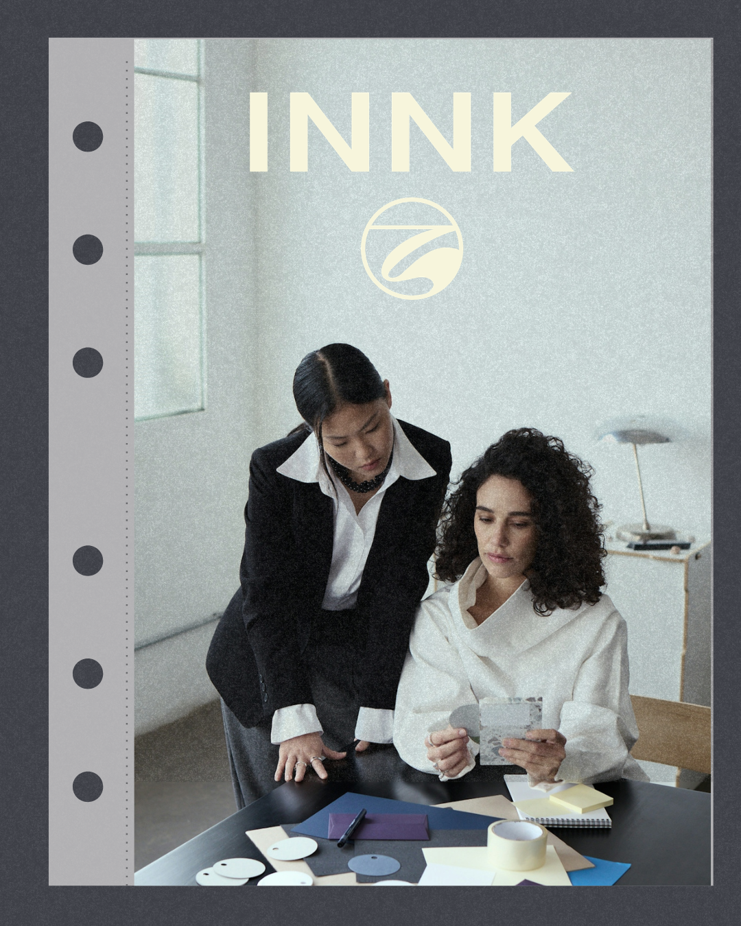 Two women collaborating at a desk with color samples, notebooks, and papers in an office. One woman is sitting and holding a photo or swatch, while the other is standing and looking at it.