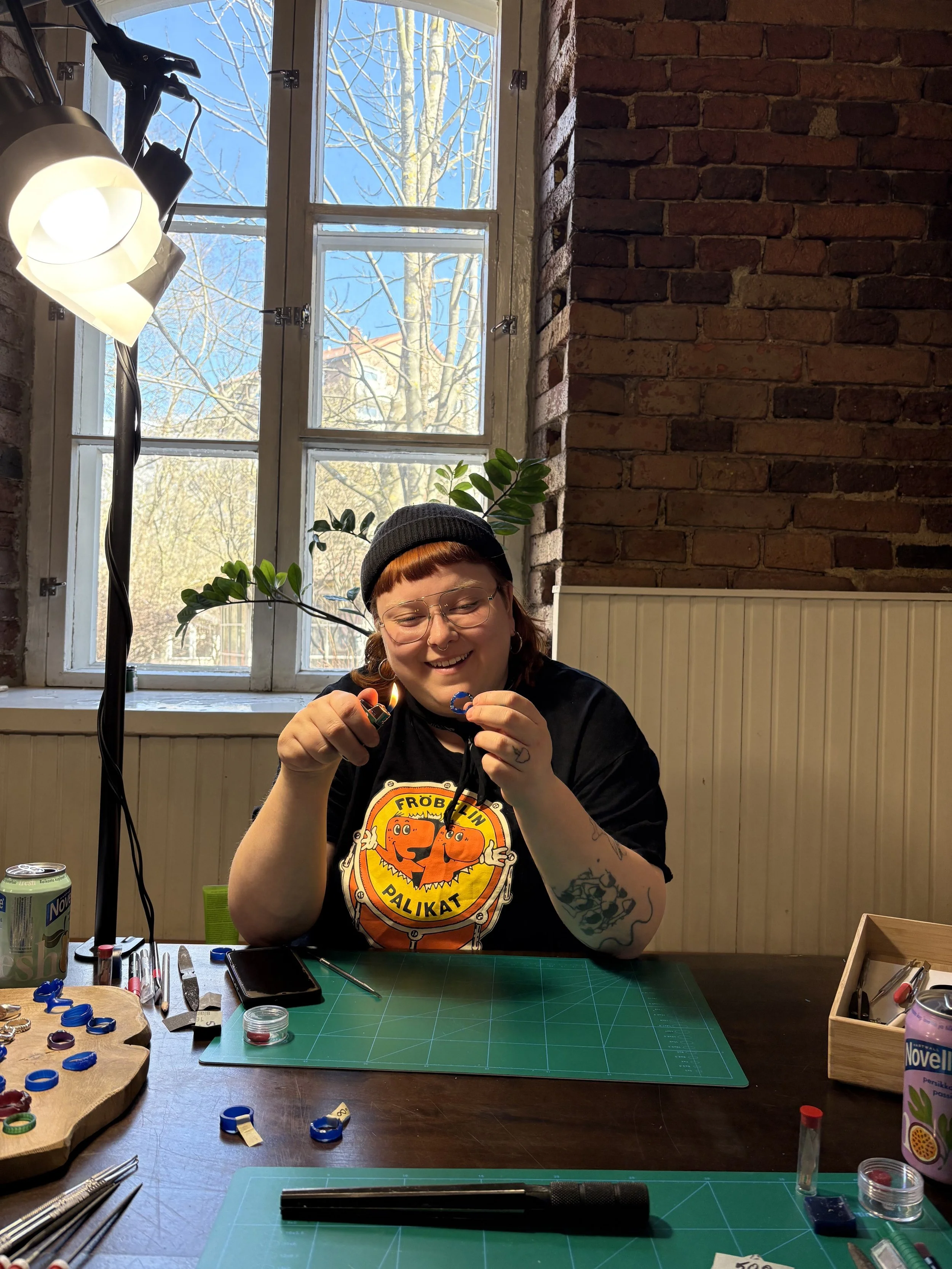 A woman with red hair and glasses smiling while working at a craft table with various tools and colorful rings, in a room with brick walls and large window showing trees outside.