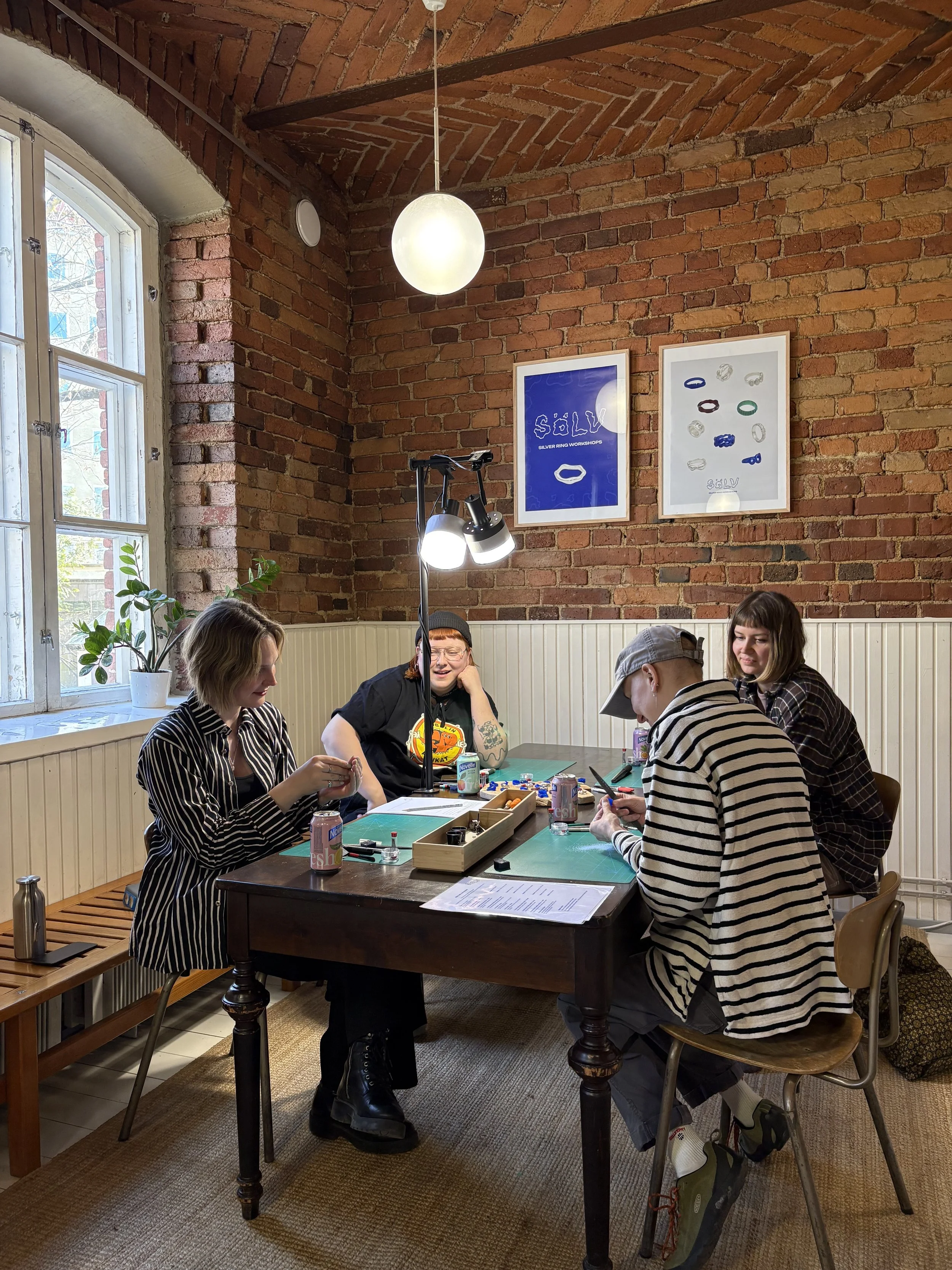 Group of five people sitting around a table playing a tabletop game in a room with brick walls and a large window, decorated with framed artwork and a potted plant.