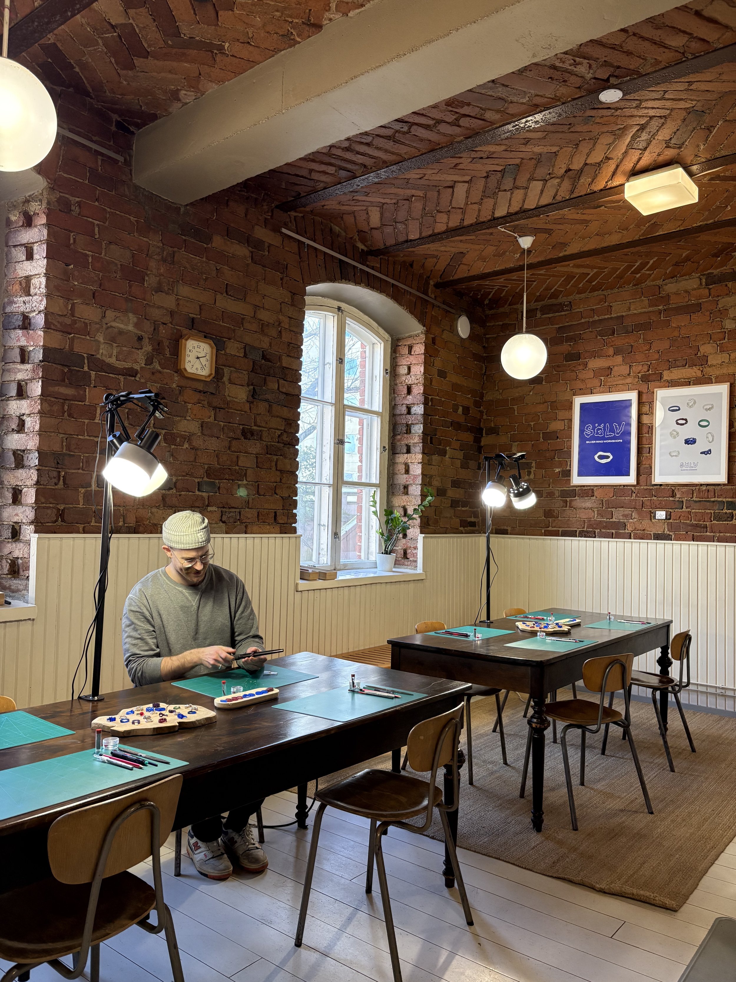 A man sitting at a wooden table in a room with brick walls and arched windows, playing a board game with game pieces on the table. There are additional tables with chairs, a carpet, framed art on the wall, and hanging ceiling lights.