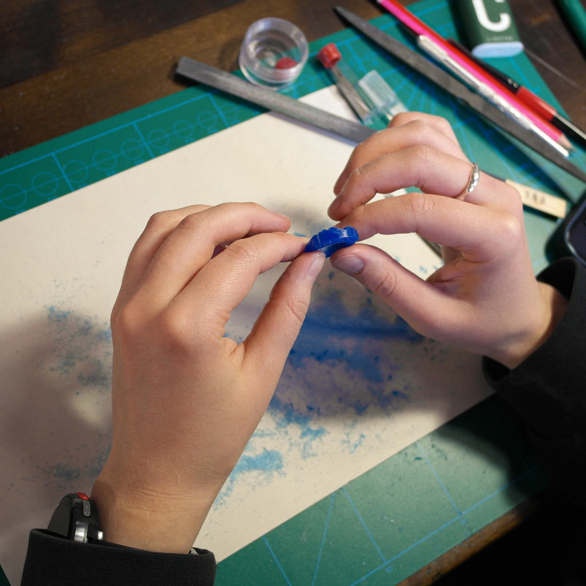 Close-up of a person's hands holding a small blue object or form, with a cutting mat, rulers, a knife, and a green object in the background on a wooden surface.
