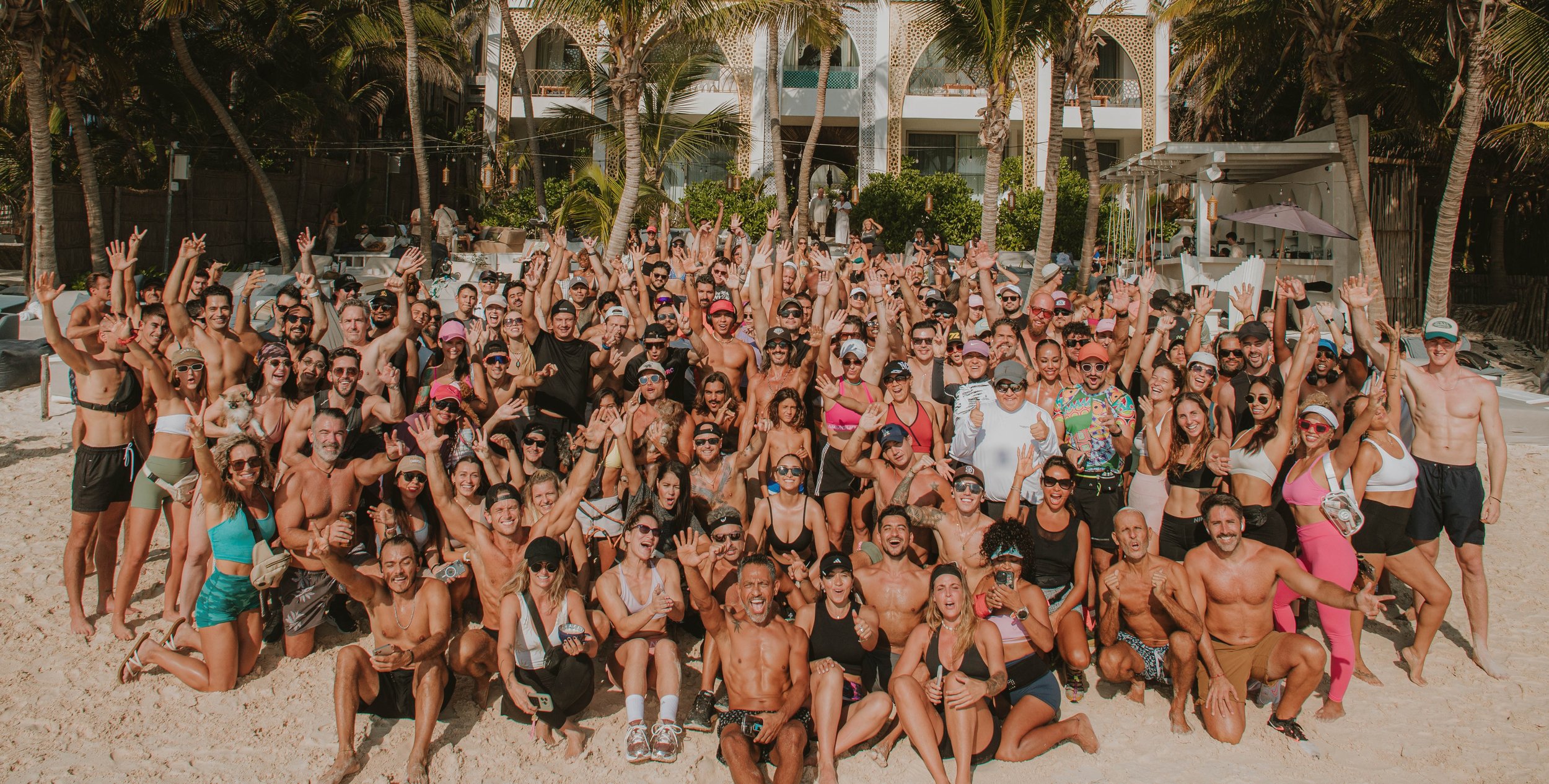 A large group of people gathered on a sandy beach, smiling and waving at the camera. Many are wearing swimsuits, sunglasses, and hats. The background shows a building with balconies, palm trees, and beachside structures, indicating a tropical or beach resort setting.