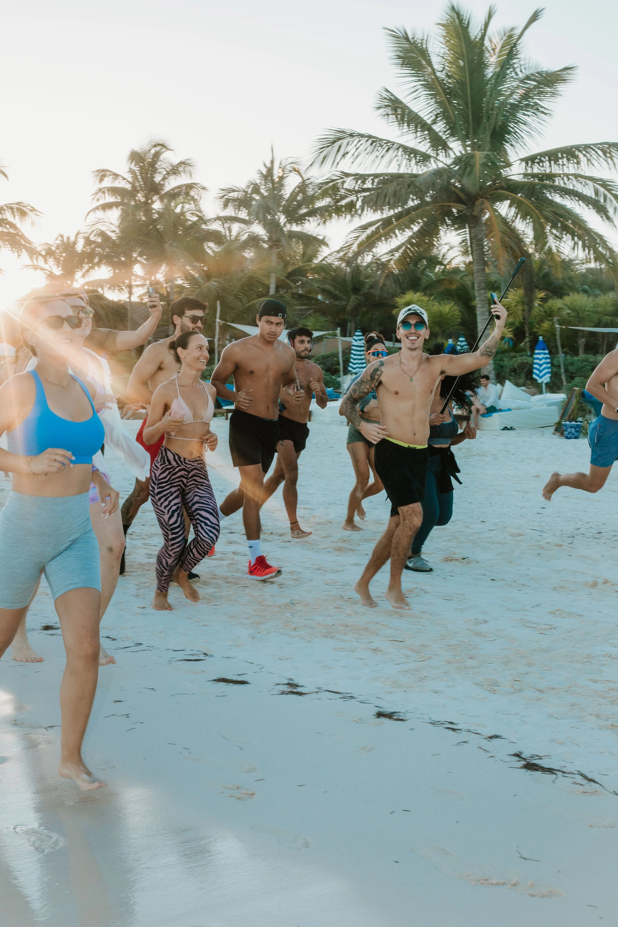 Run Tulum - Group of people running on the beach with palm trees and blue umbrellas in the background, during sunset.