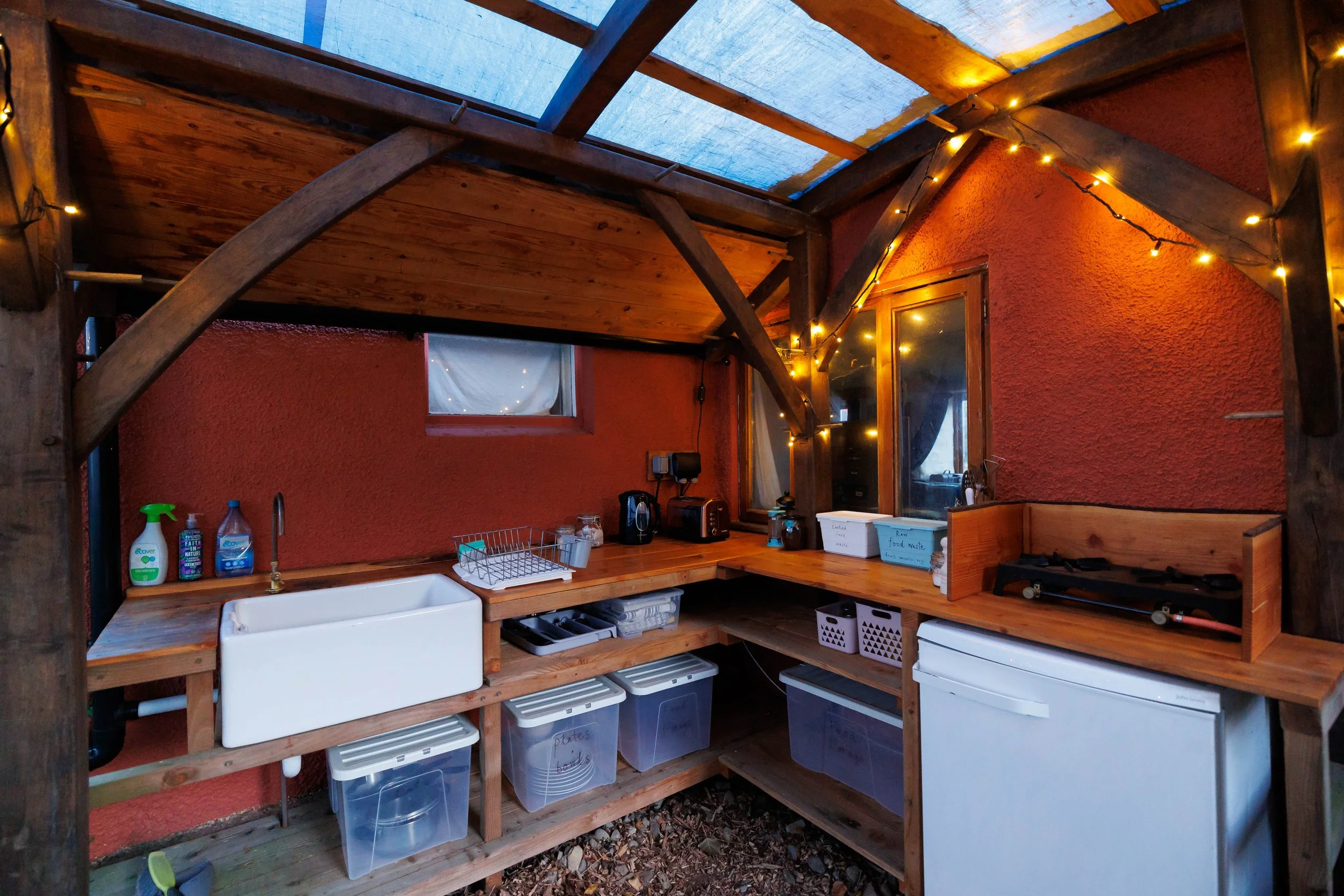A rustic outdoor kitchen with a wooden countertop, a white sink, and various kitchen appliances, under a roof made of wood and clear panels, decorated with string lights.