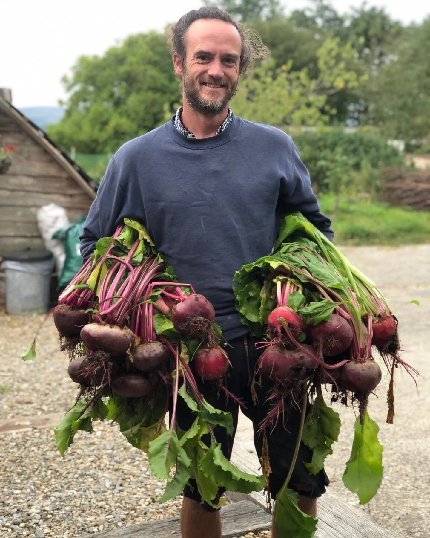 A man standing outdoors holding two bunches of freshly harvested beets with leaves attached.