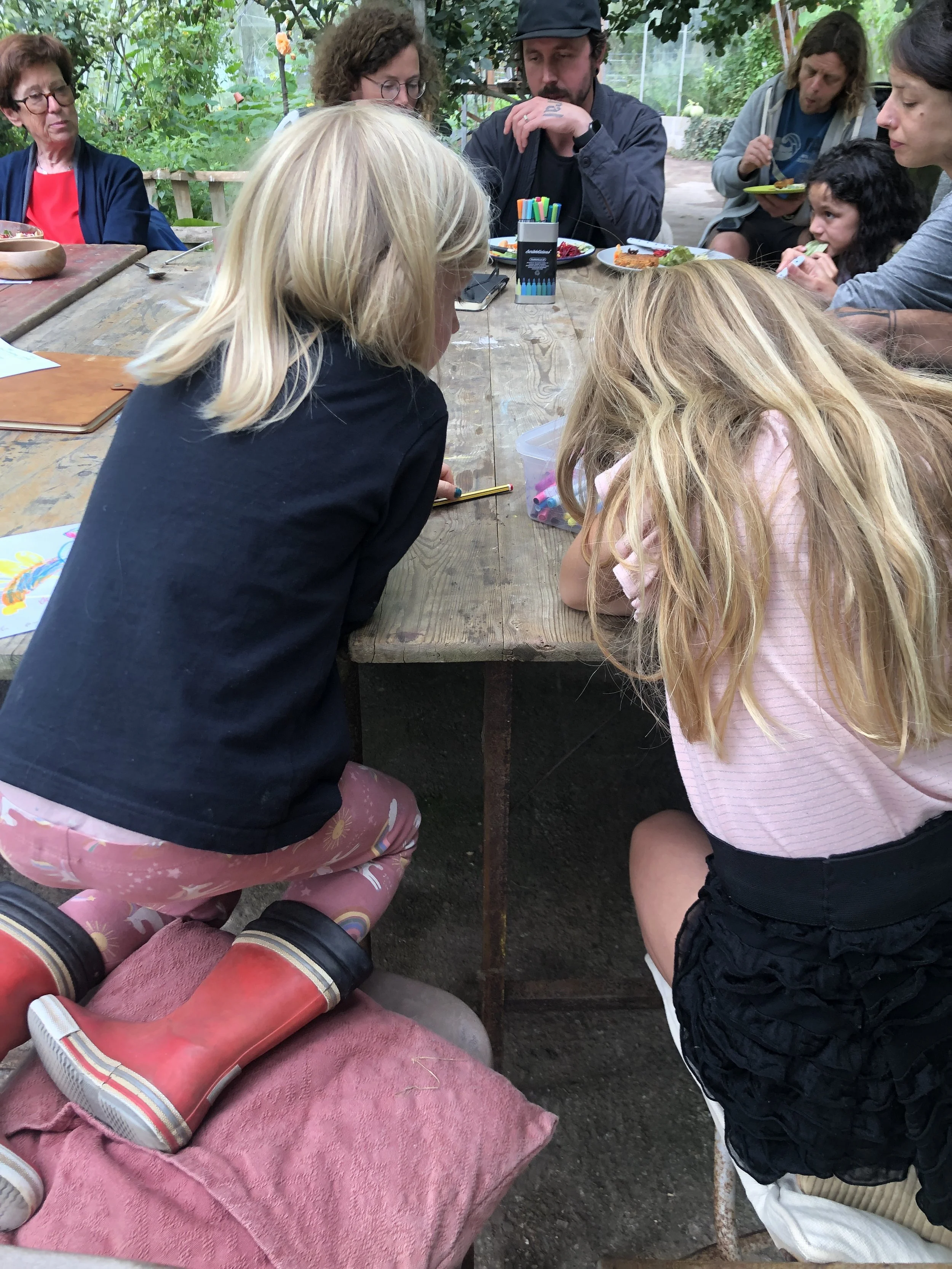 Children and adults sit around a wooden table outdoors, engaged in an activity, with art supplies and colorful markers on the table, in a lush garden setting.