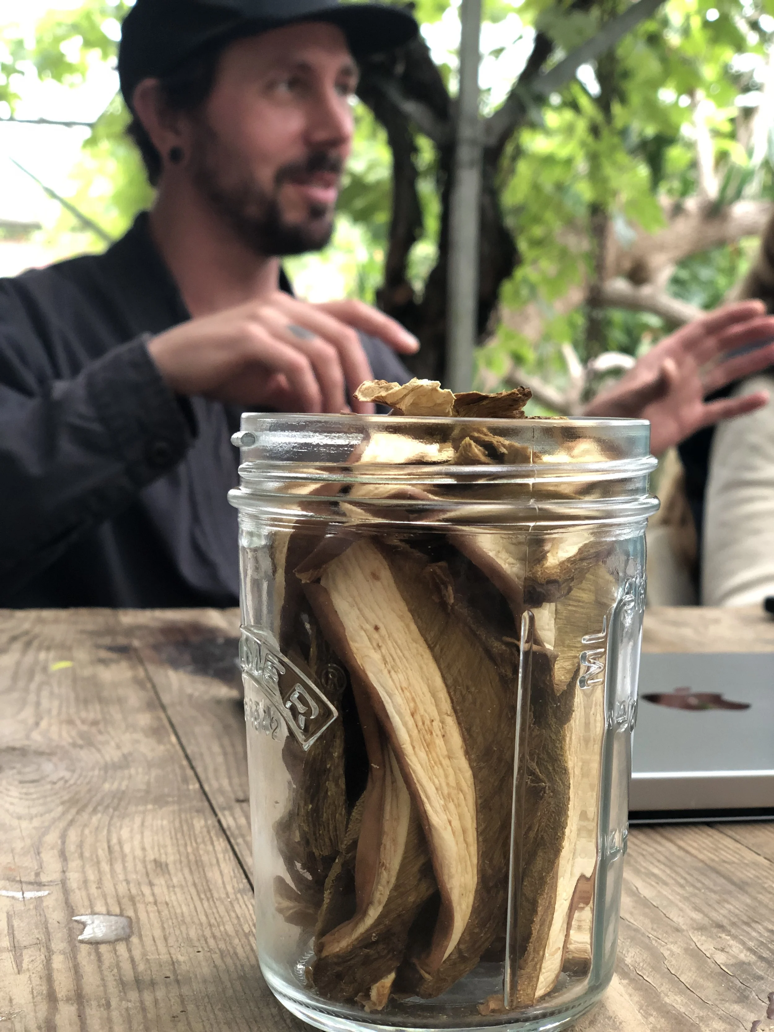 Close-up of dried mushrooms in a glass jar on a wooden table, with a blurred person in background.
