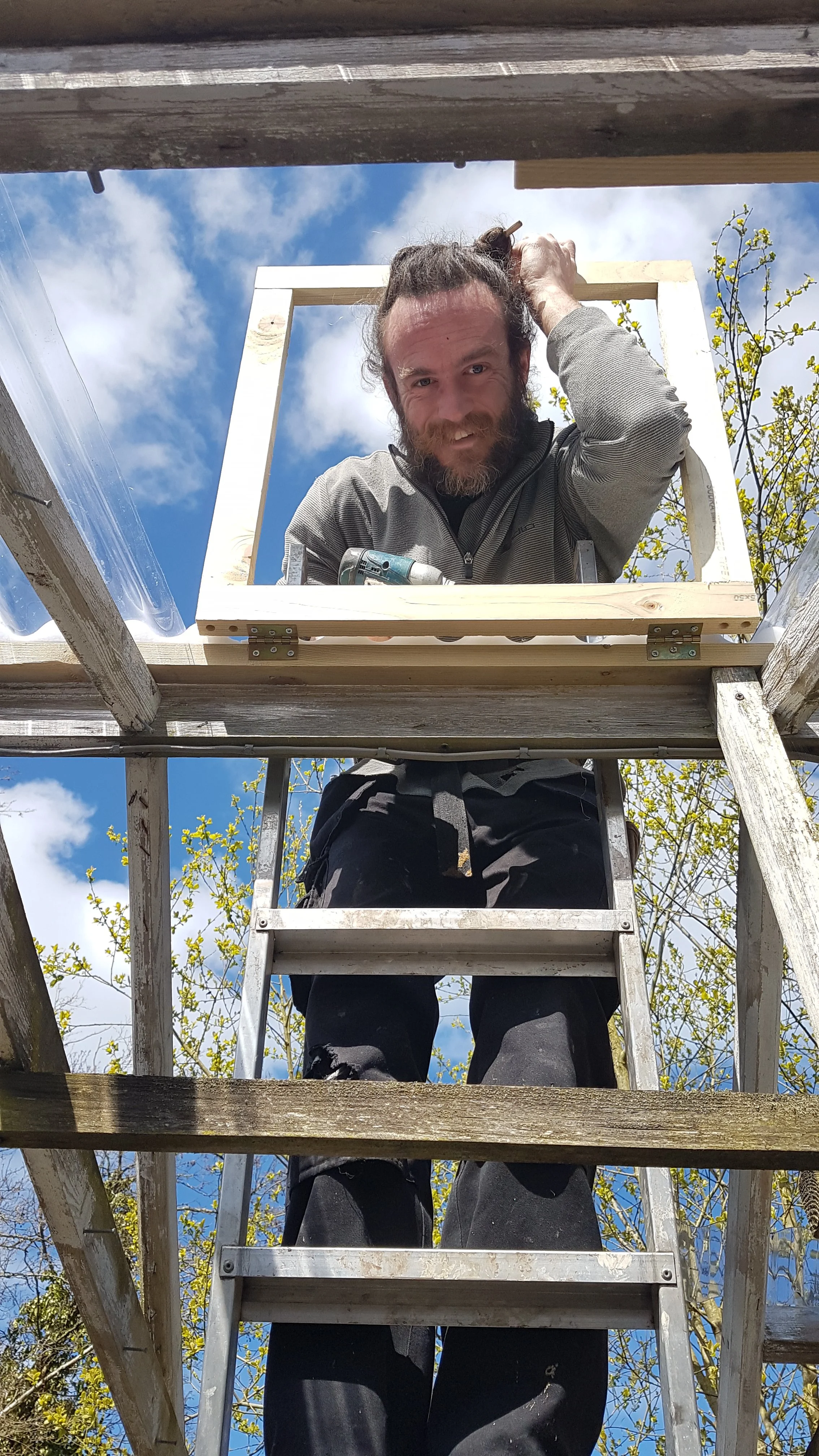 A man working on a wooden ladder outside, with a blue sky and clouds behind him, using a power drill.
