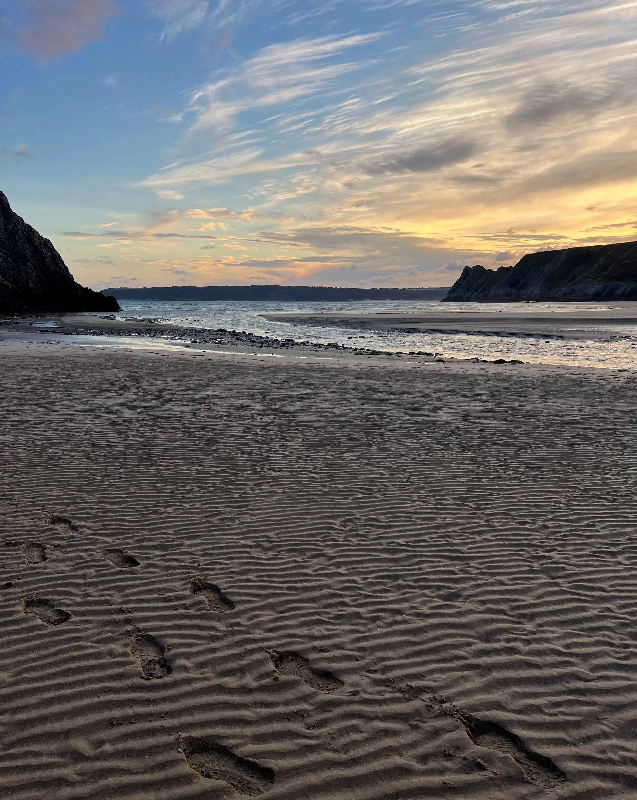 Footprints in the sand on a beach with calm water, cliffs on either side, and a sunset sky with clouds.