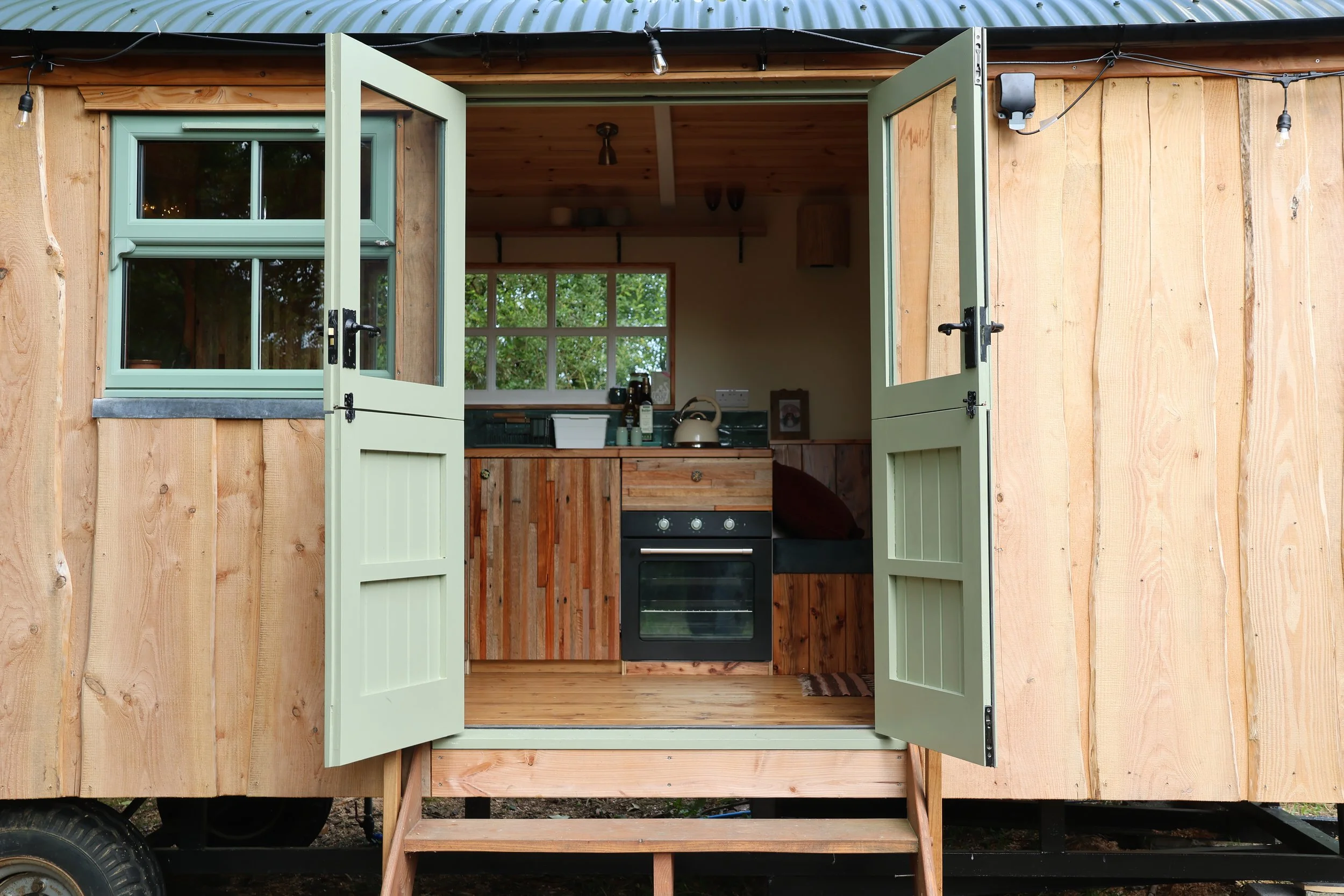 Small wooden house with open double doors revealing a rustic kitchen with wooden cabinets, a stove, kettle, and a window showing greenery outside.