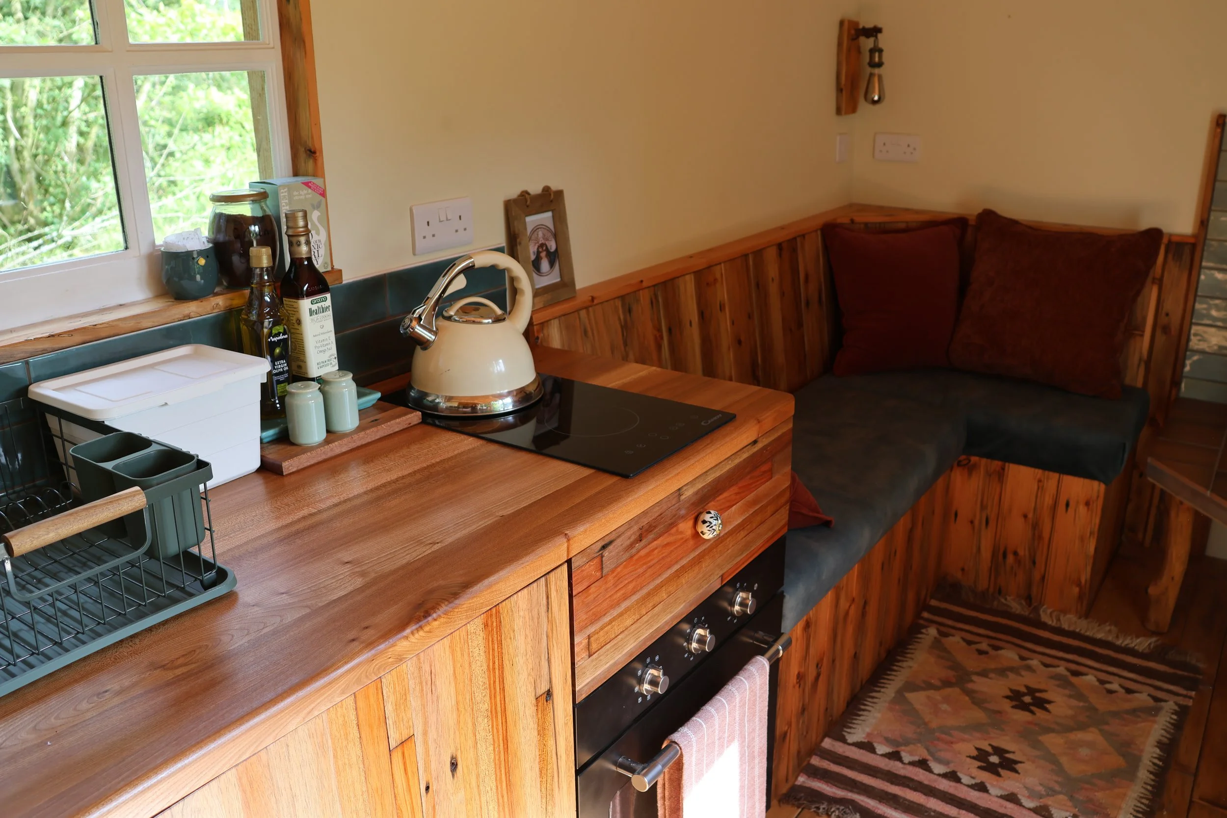 A cozy rustic kitchen corner with a wooden countertop, a stovetop with a cream-colored kettle, and a small built-in seating area with cushioned benches and pillows. There is a window showing green foliage outside.