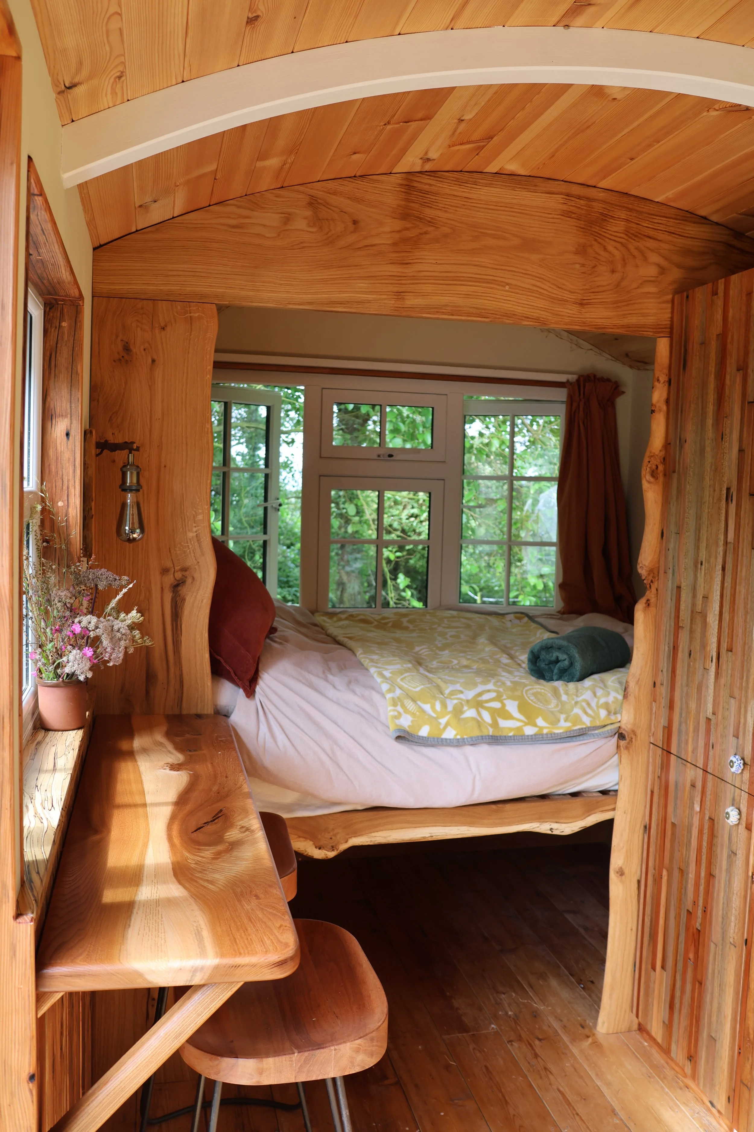 Small wooden cabin bedroom with a bed against the windows, a wooden desk and stool, and natural wood paneling throughout.