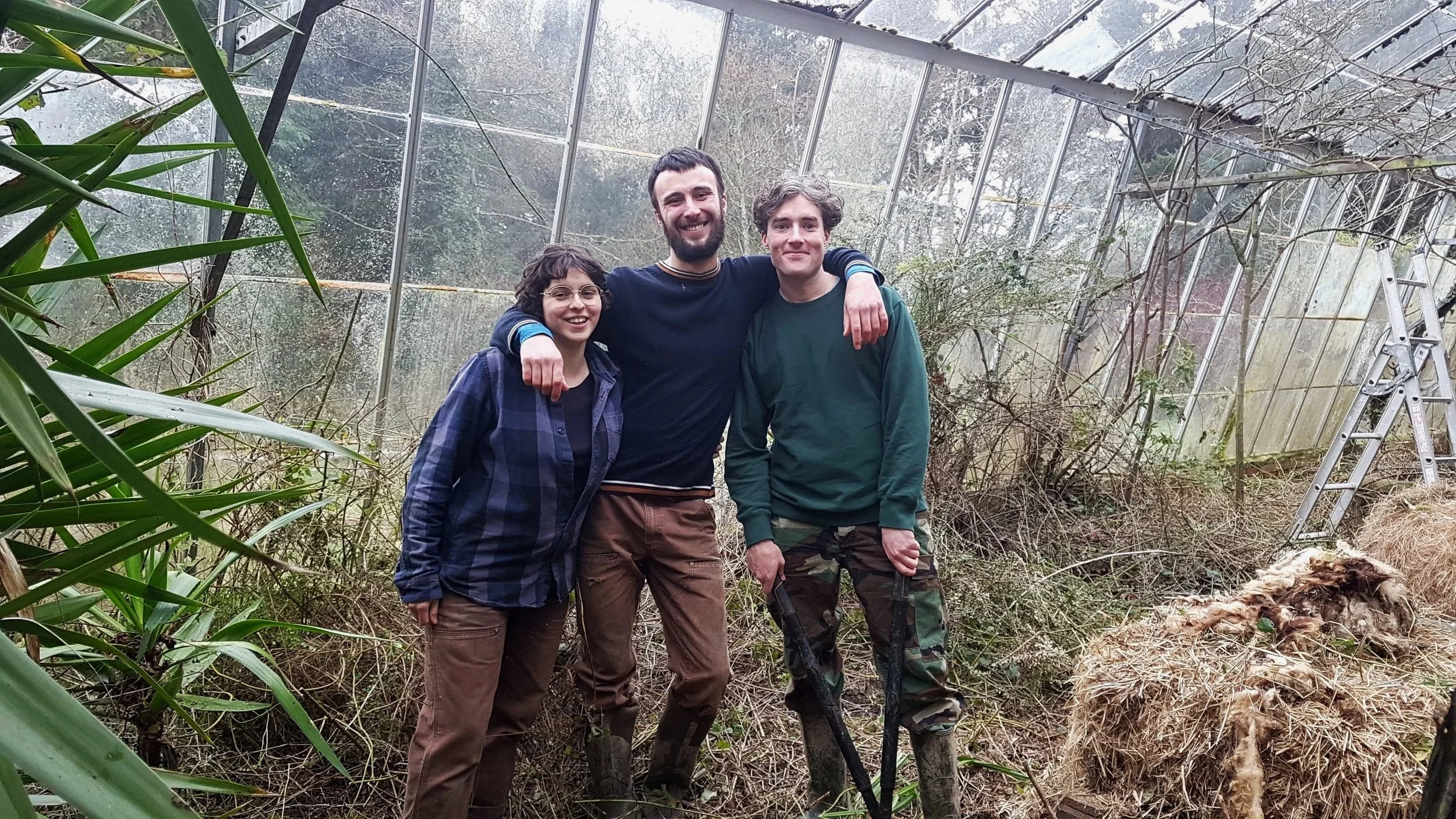 Three smiling people standing in a greenhouse with overgrown plants and a ladder on the right.