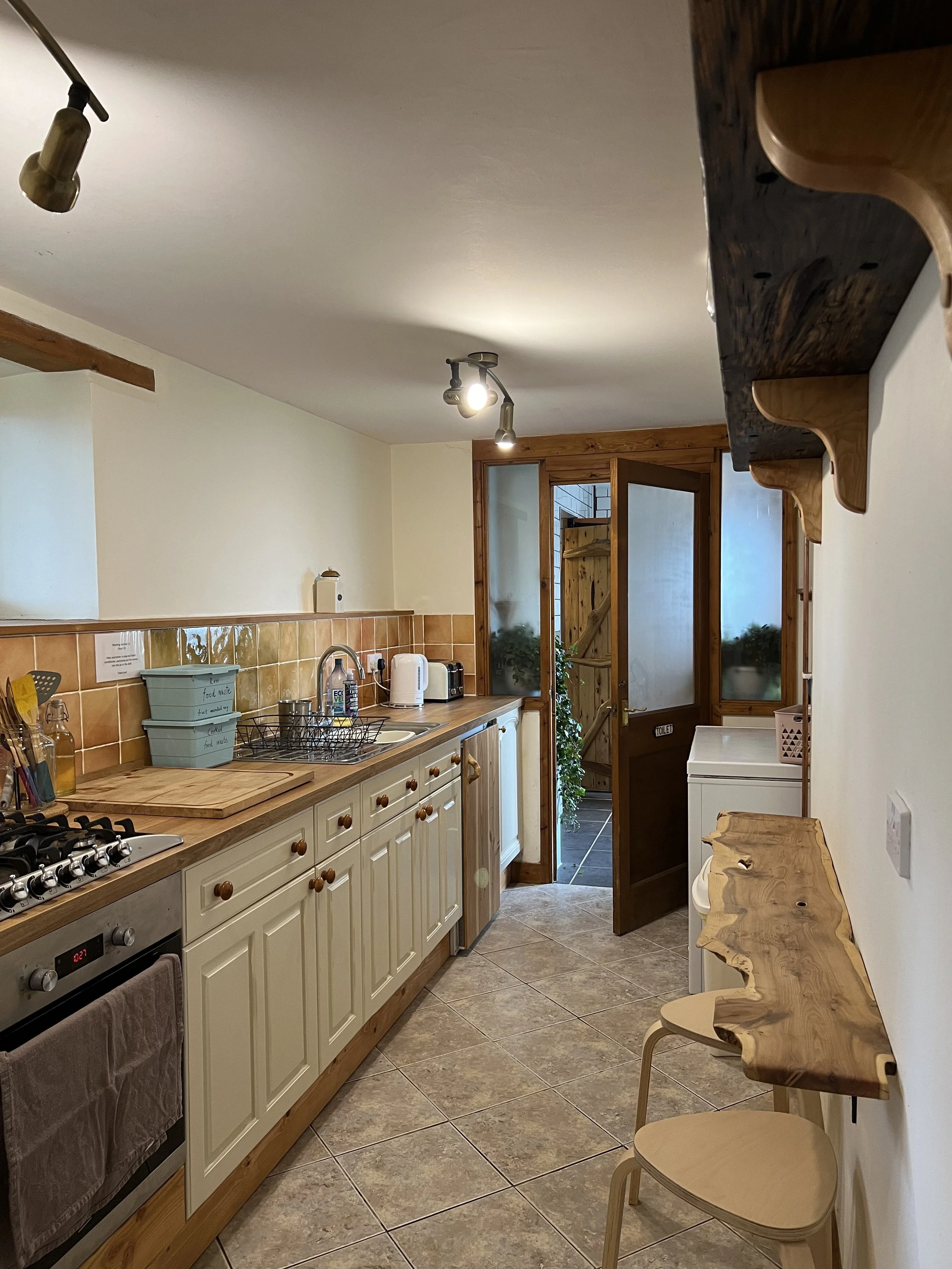 A cozy kitchen with cream-colored cabinets, a wooden countertop, and brown tiled backsplash. There are various appliances including a stove, kettle, and toaster. A small wooden shelf and two stools are attached to the right wall. A door at the back leads to an outdoor area with plants.