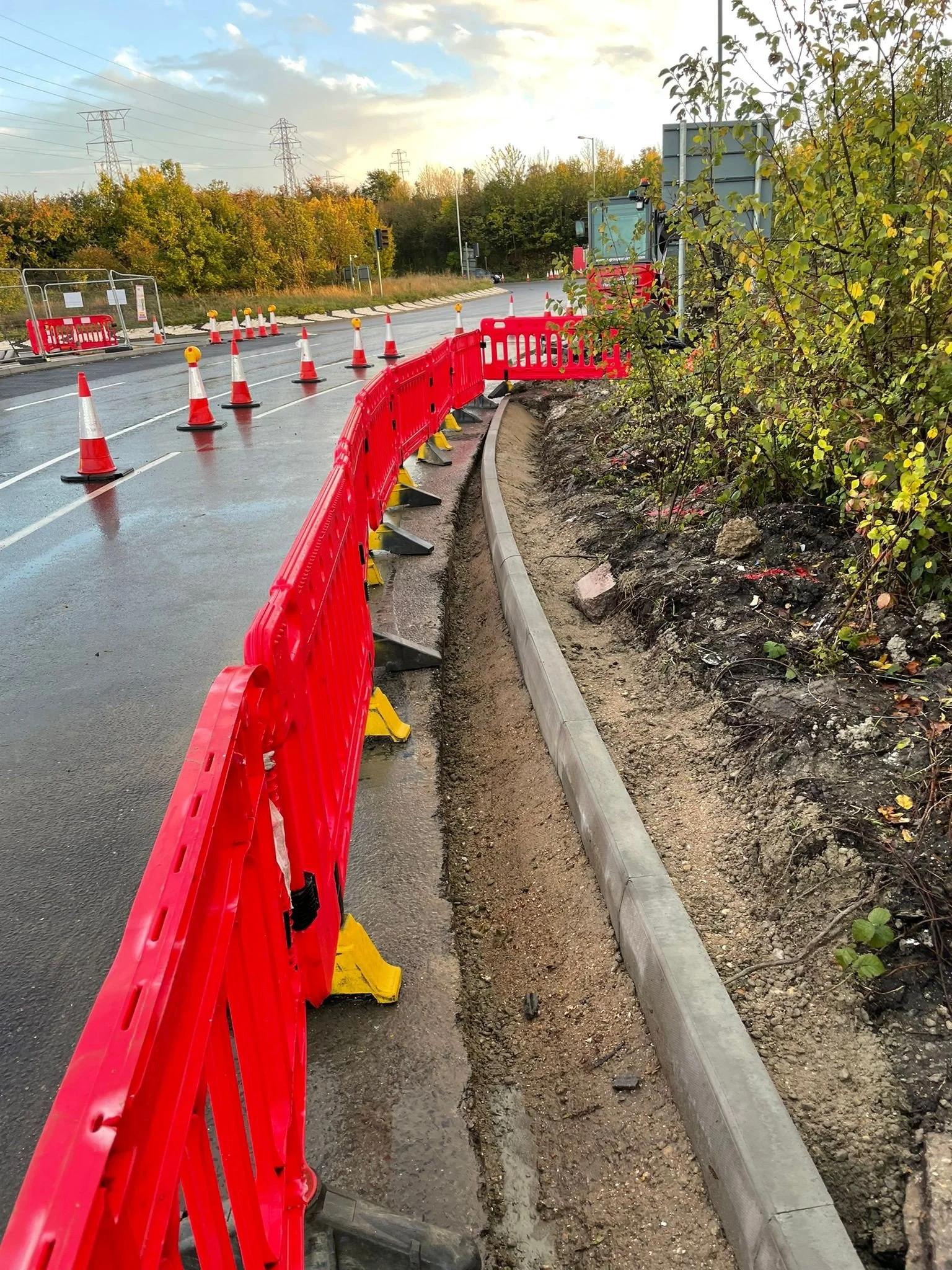 Construction site on a road with orange safety cones and barriers, and a curved sidewalk with bushes on the side.