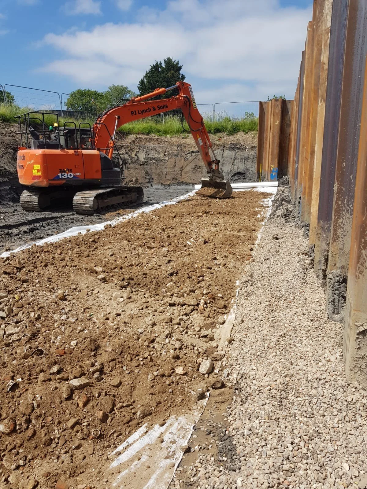 Construction site with an orange excavator digging and moving dirt along a retaining wall with wooden panels, under a partly cloudy sky.