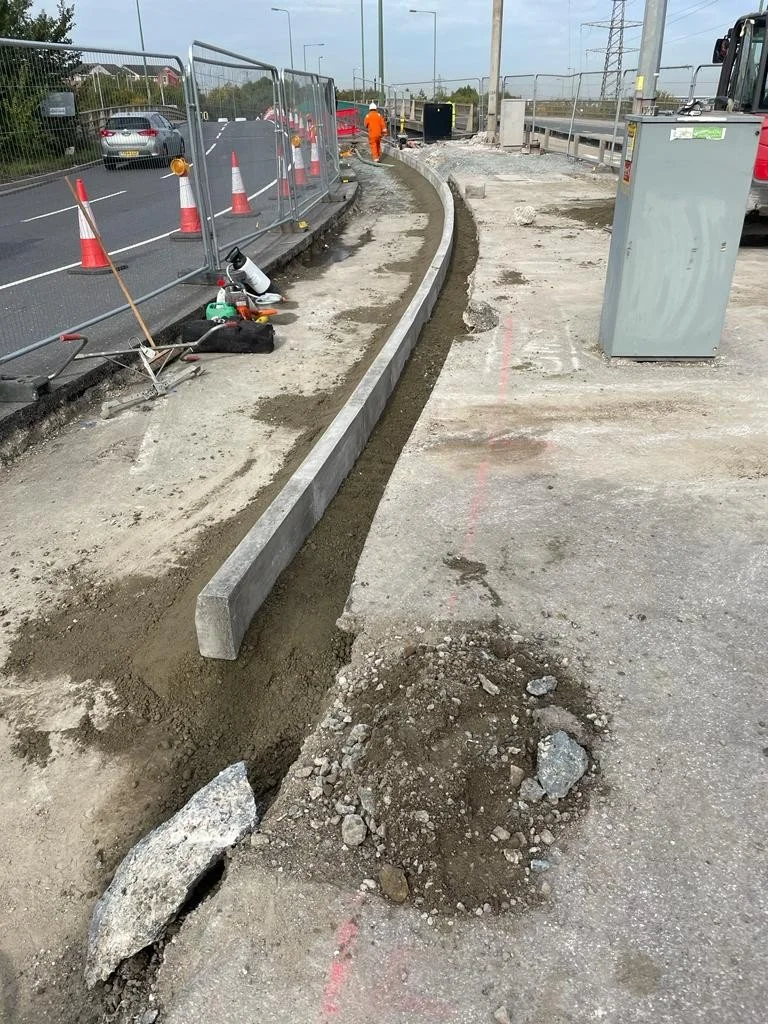 Construction workers working on sidewalk curb installation at a roadside, with traffic cones and equipment nearby.