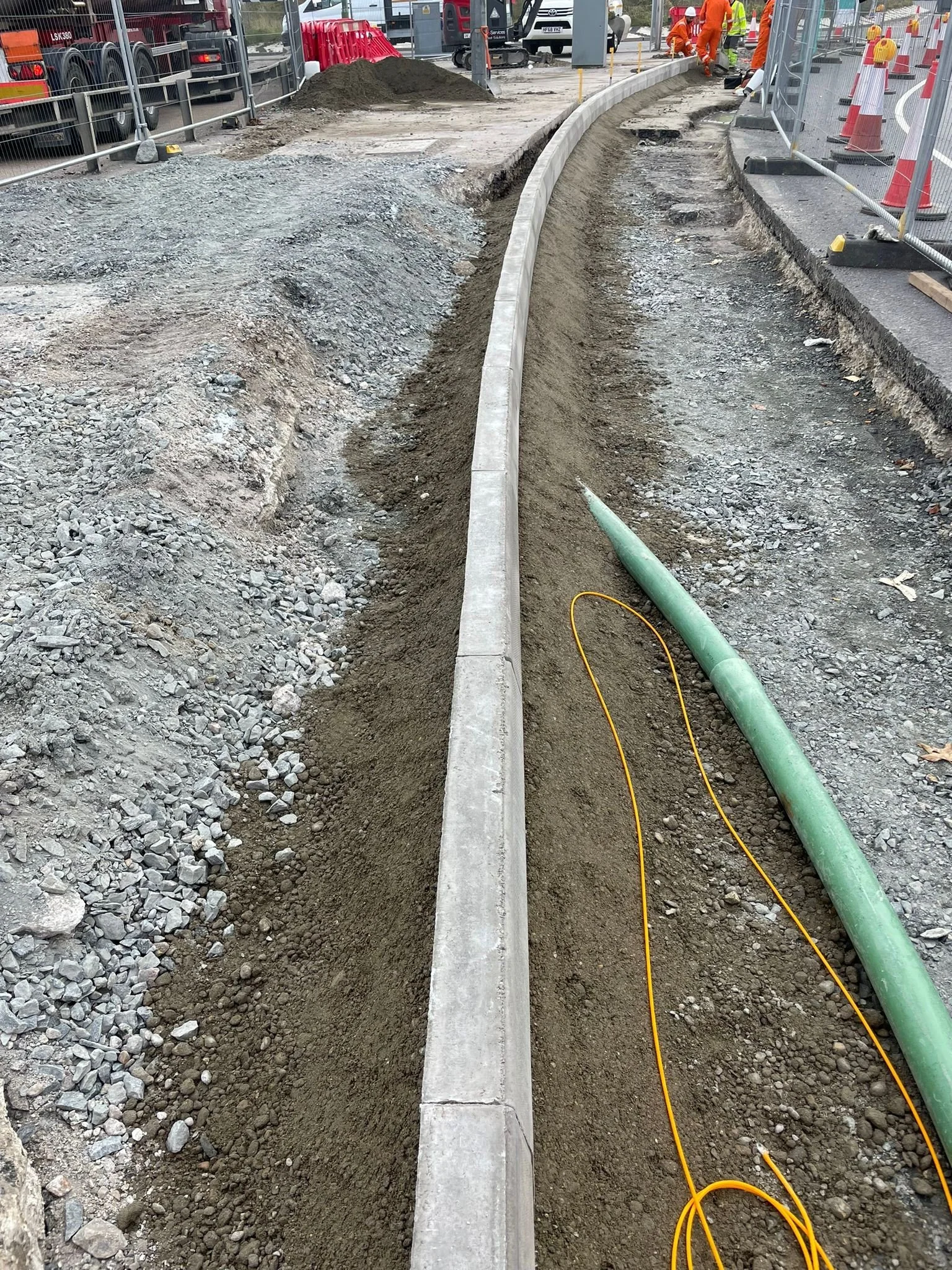 Construction site where workers are building a sidewalk with concrete curbing along a curved pathway, with construction barriers, safety cones, and equipment nearby.