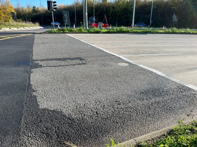 A paved road transition to a parking lot with a traffic light in the distance. The road surface appears to be recently repaved or under construction.