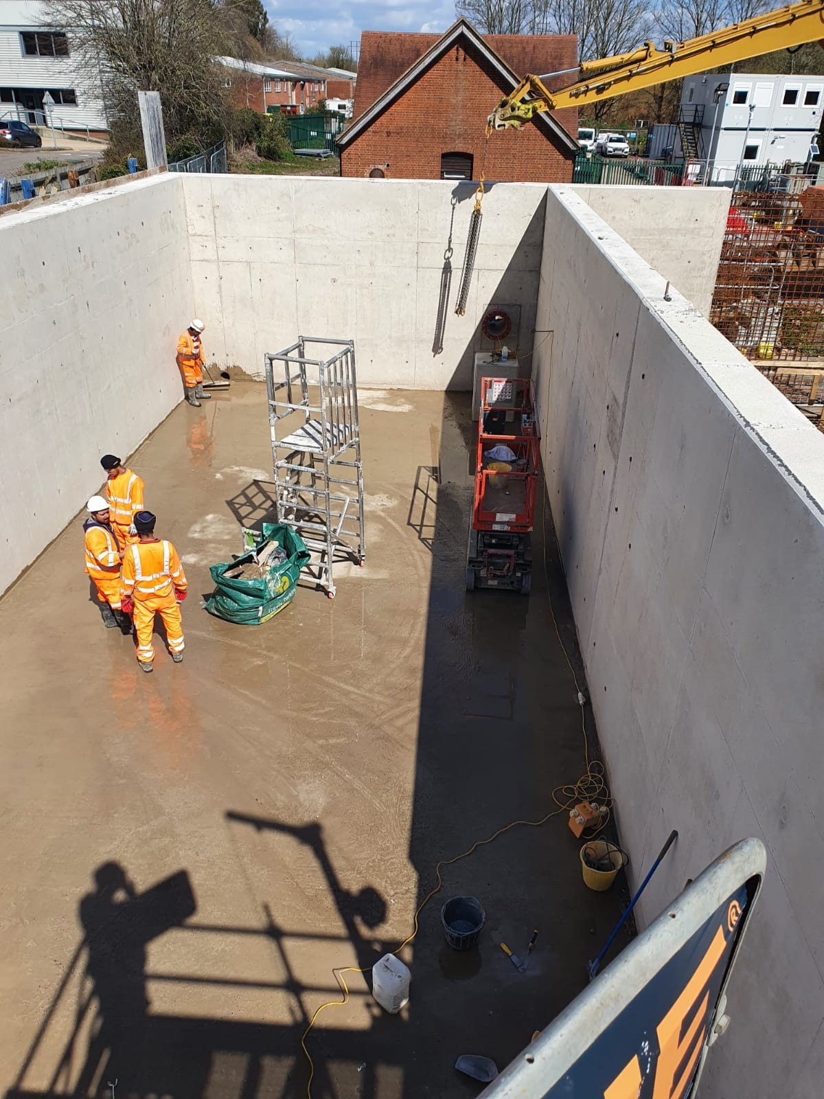 Construction workers in orange safety vests and helmets working inside a large concrete basin, with construction equipment and tools around. A crane lifts a heavy object overhead, casting shadows on the ground.