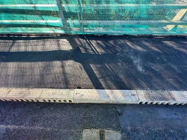 Close-up of a city sidewalk with a metal drainage grate and shadows from a glass railing or fence.