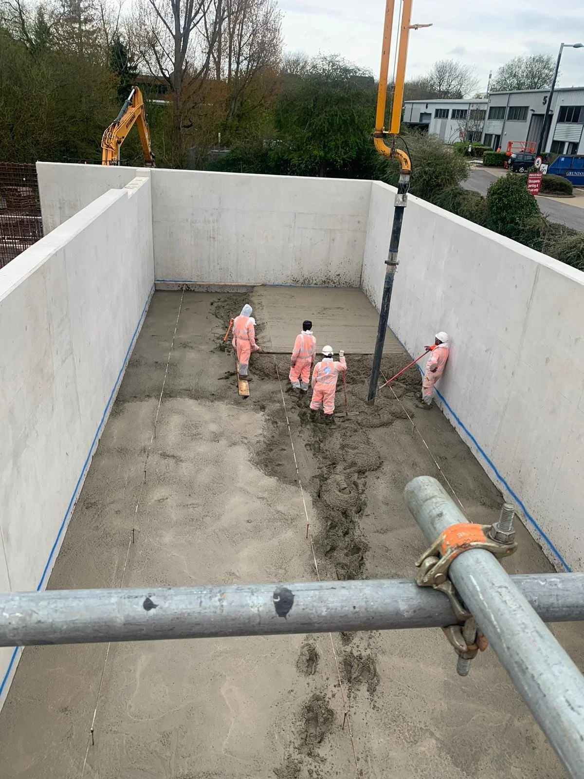 Construction workers pouring concrete inside a foundation wall, with concrete pump and construction equipment visible.