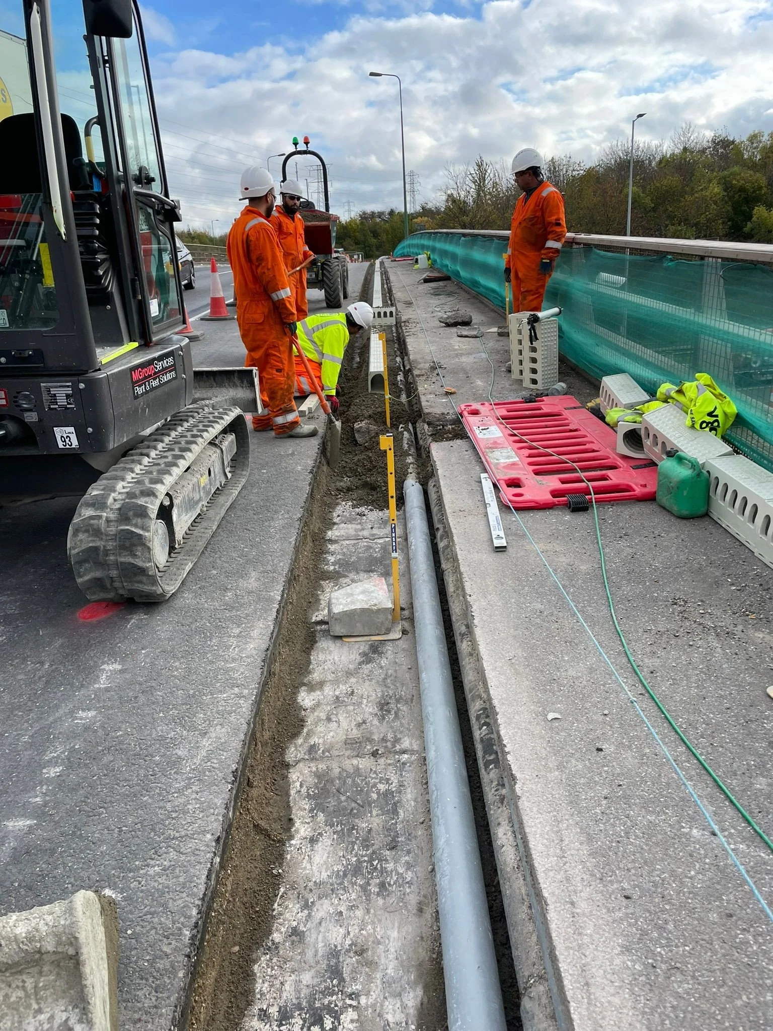 Construction workers in safety gear working on a roadside project, installing underground pipes, with equipment and barriers around the site.