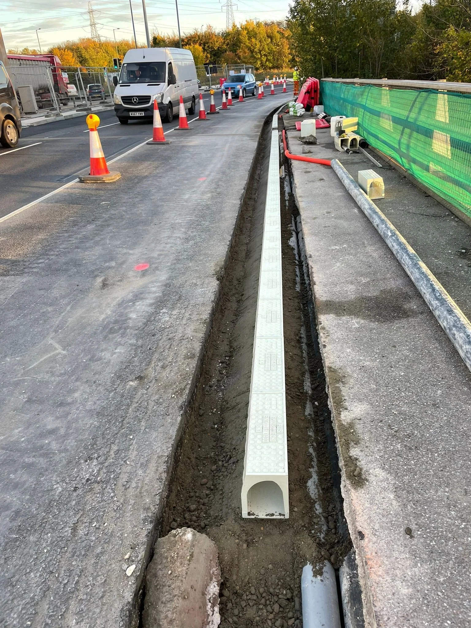 Road construction site with orange traffic cones, a white van, and ongoing utility work along the side of the road.