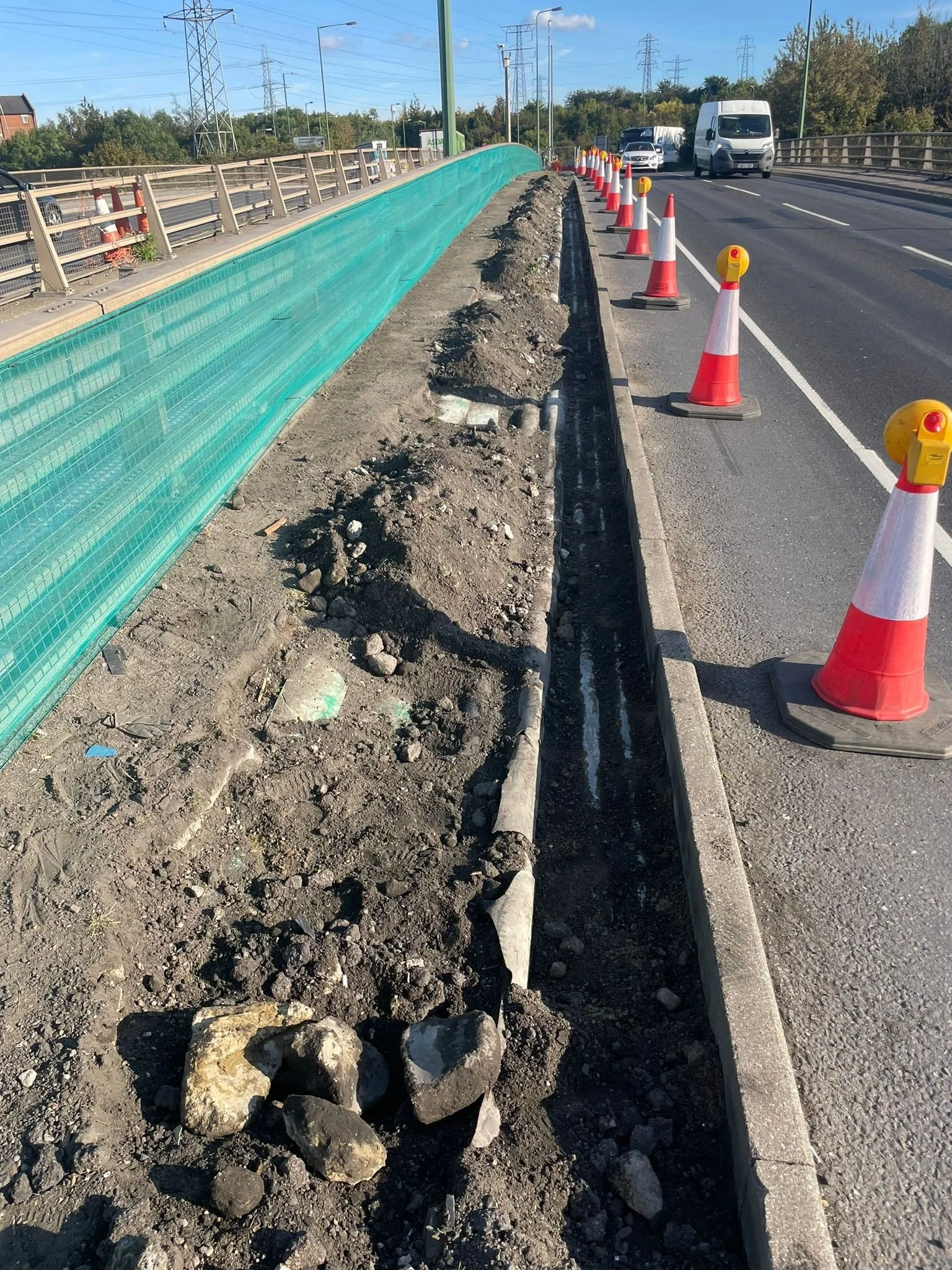 Road construction with orange traffic cones along a divided highway, partially excavated sidewalk with exposed pipes and rocks, green protective fencing, and vehicles in the background.
